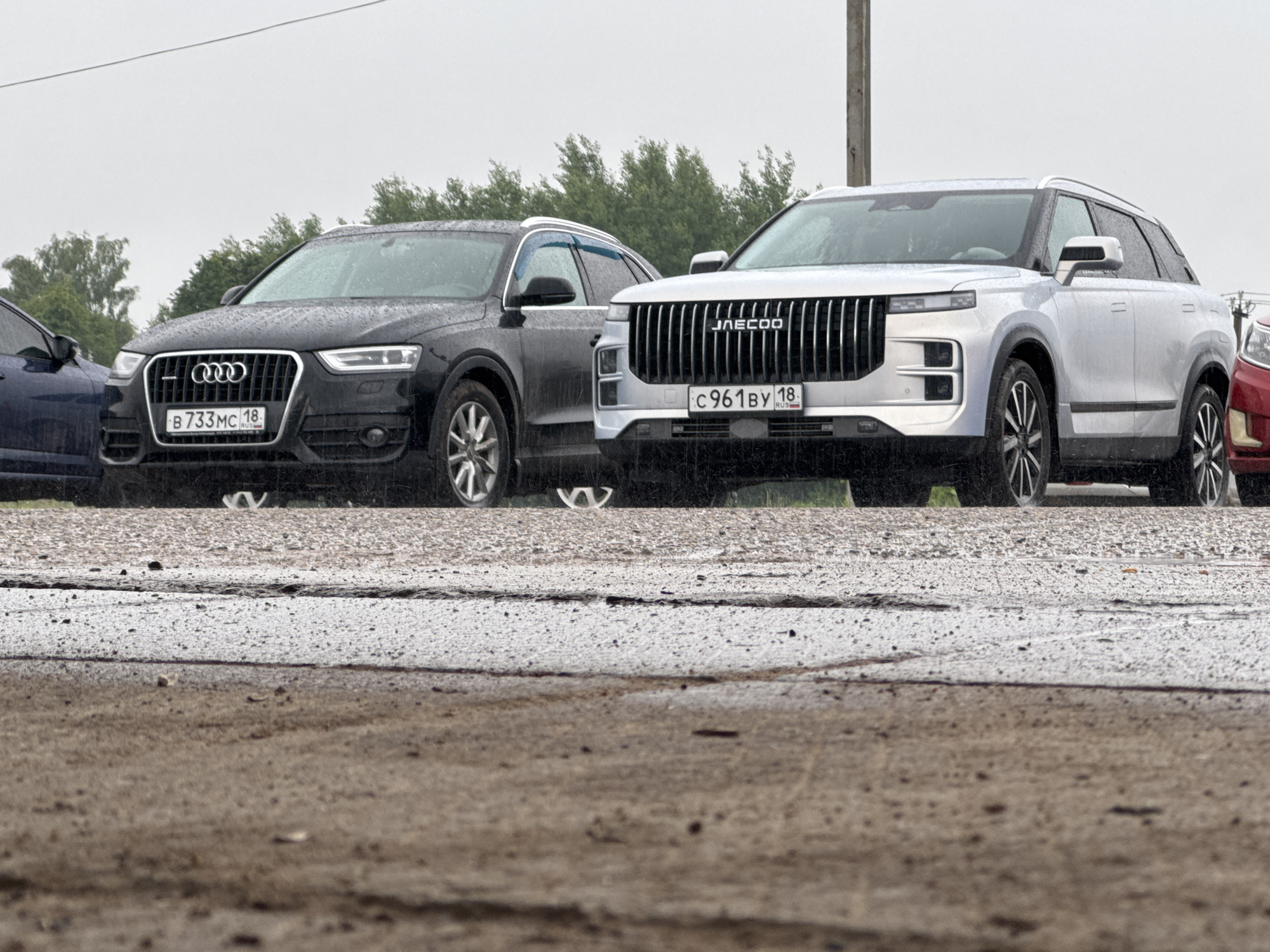 The image captures a scene in Mozhga, Russia, featuring two prominent parked SUVs under an overcast sky, with evidence of recent or ongoing rain.
In the foreground, a black Audi Q3 is parked on the left, viewed from its front-left quarter. Its grille features the Audi rings logo, and its Russian license plate, "B 733 MC 18 RUS," is clearly visible. To its right, a silver JAECOO SUV is parked, also viewed from the front-left. Its distinctive grille features vertical slats and prominently displays the "JAECOO" brand name. Below it, the Russian license plate "C 961 BY 18 RUS" is visible. Both vehicles, and others partially seen, are covered in numerous water droplets, indicating rain. A dark blue car is partially visible on the far left, next to the Audi, and a red car is partially visible on the far right, next to the JAECOO.
The setting appears to be an outdoor parking area or roadside. The ground in the foreground is a textured, cracked asphalt or concrete surface, appearing wet and speckled, consistent with rain. In the background, blurred green trees are visible under a uniformly grey and dull sky. A utility pole stands vertically behind the silver JAECOO SUV. The overall atmosphere suggests a quiet, damp daytime scene.
