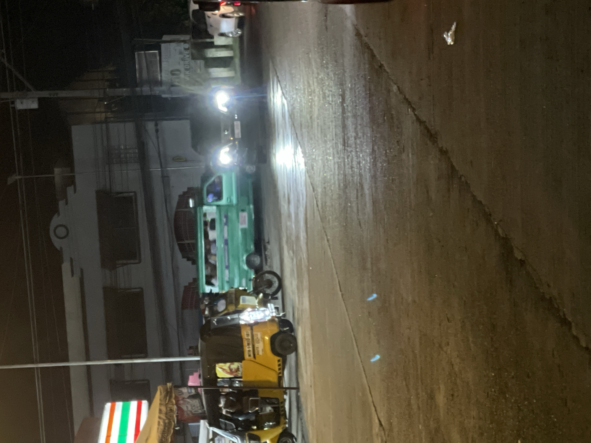 At night, on a paved road in Davao City, Philippines, several vehicles are visible. In the foreground, a yellow motorized tricycle with a black canopy is parked. To the left of the tricycle, a portion of a 7-Eleven store is illuminated. Further back, a green passenger jeepney is driving on the road. In the center of the image, the headlights of an oncoming car are intensely bright. In the background, buildings line the street, and utility poles with wires are present. The road surface appears wet and reflective, indicating recent rain or moisture.