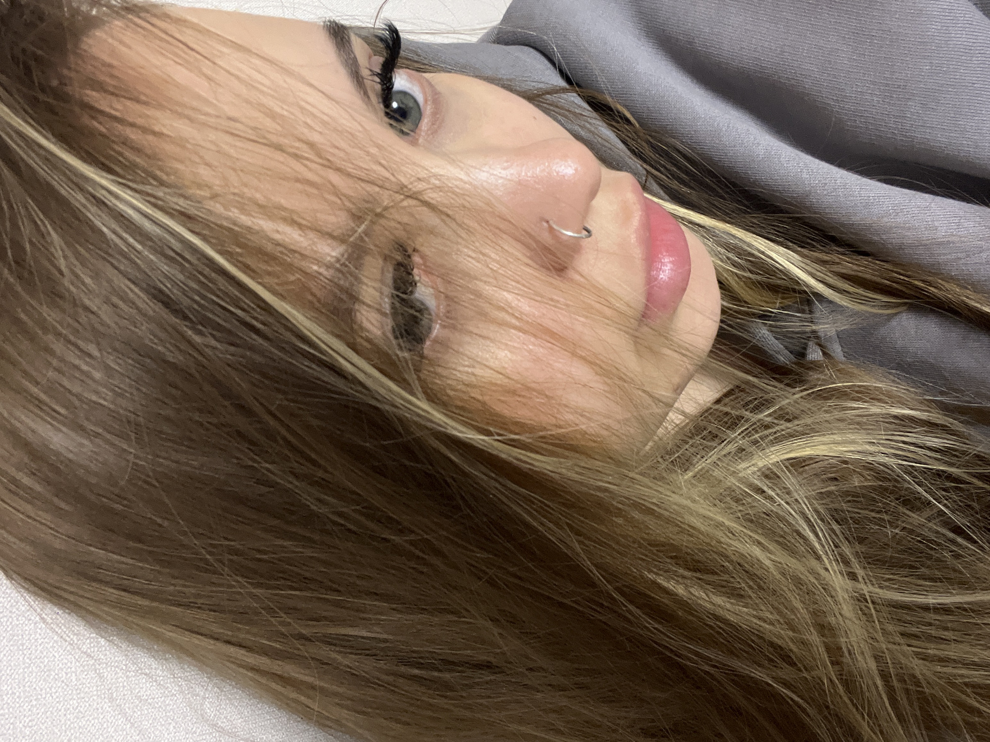 A close-up, eye-level shot shows the upper body of a young woman lying down and looking upwards. Her head is tilted to her left, with her right eye, nose, and mouth prominently featured. Her gaze is directed towards the top right of the frame.
Her right eye has a light-colored iris, possibly blue or green, with long, dark eyelashes. Her nose features a small, silver hoop piercing on the right nostril. Her lips are slightly parted, glossy, and pink. Long, multi-toned hair, appearing to be light brown with lighter blonde or streaked highlights, falls across her face, partially obscuring her left eye and chin, and spreads around her head. A grey, textured fabric, likely a sweatshirt or hooded top, is visible on the upper right side of the image. The background consists of a plain, light-colored surface, suggesting an indoor setting. The lighting is soft, highlighting her facial features and hair. Her expression appears calm and neutral. No text is visible in the image. The location is Krasnodar, Russia.