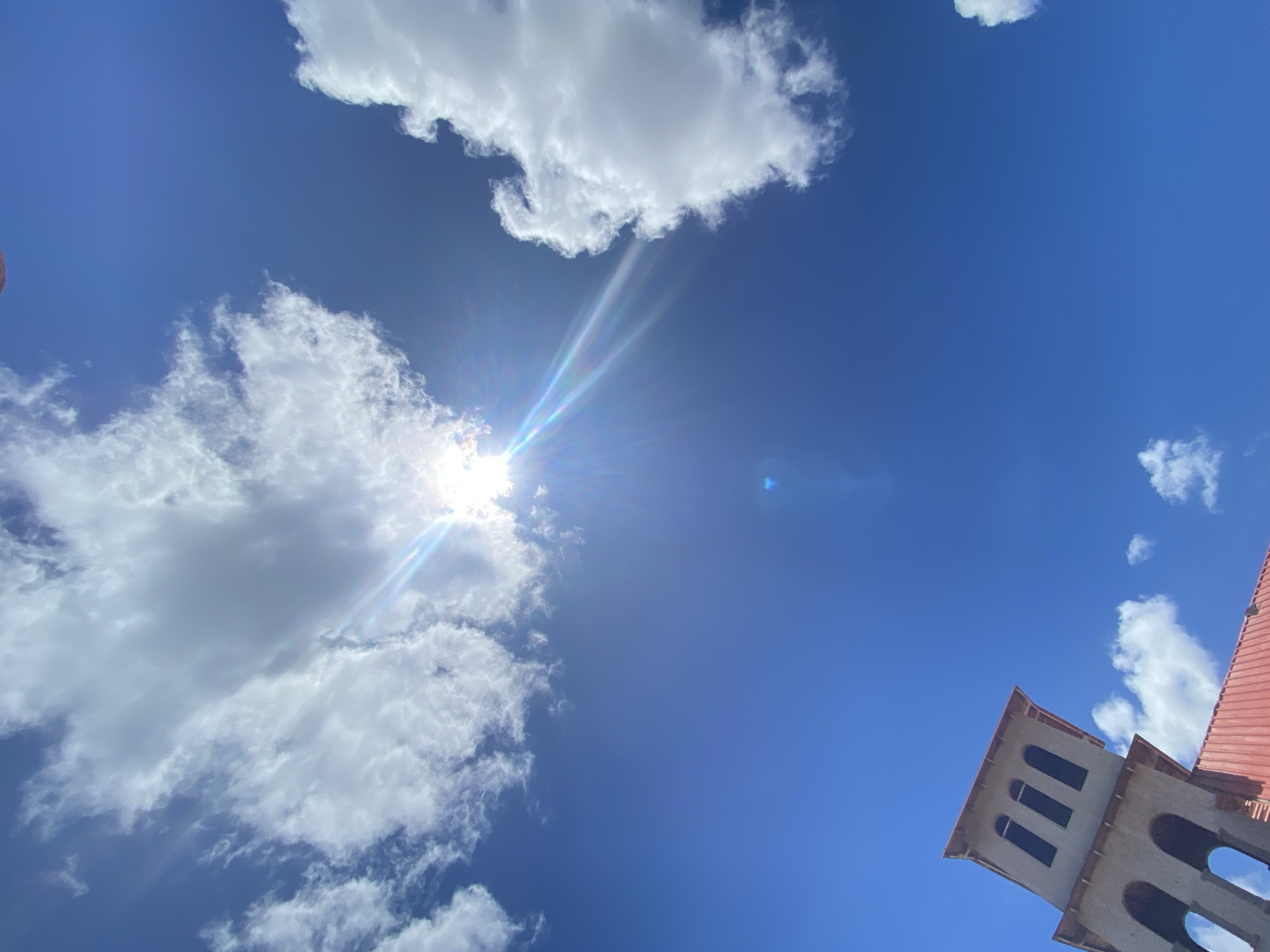 The image is a view of the sky looking up, with white clouds scattered across a bright blue background. The sun is partially obscured by clouds on the left, creating a lens flare effect. In the lower right corner is a section of a tower with arched windows and a red tile roof.
The location of this image is most likely Juan Griego, Venezuela based on the details of the architectural design, particularly the towers, that are a feature of local historic sites.