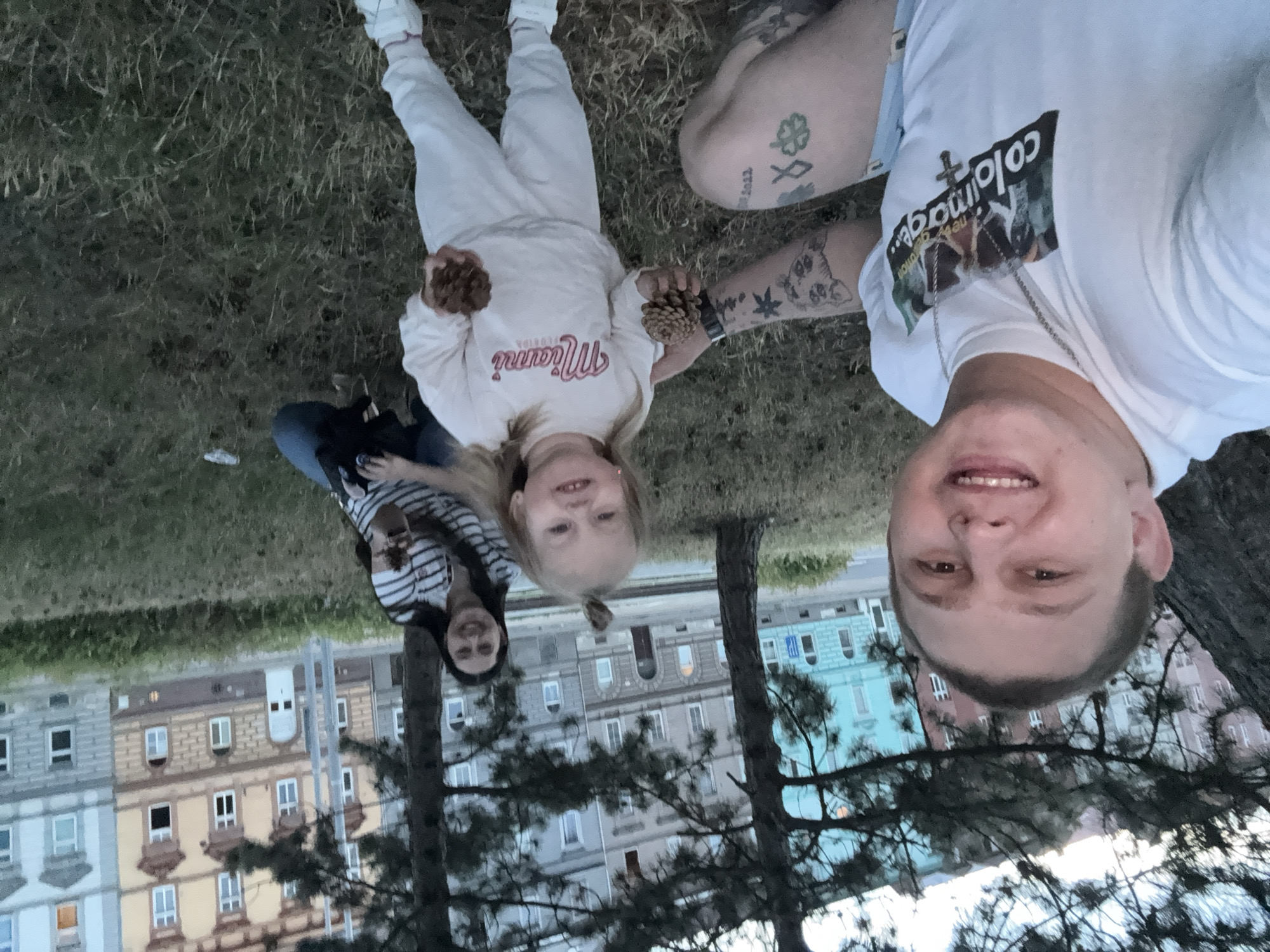 This is a close-up, slightly blurred, selfie-style photograph taken outdoors in Pilsen, Czechia, during daylight hours. The foreground is dominated by a smiling man on the left, wearing a white t-shirt with a graphic print and a necklace. His left arm, visible to the right, is tattooed. Next to him and slightly behind is a young child, likely a girl, wearing a light-colored tracksuit with "Miami" printed on it. She is holding pinecones in both hands. In the background, slightly out of focus, a woman in a striped shirt is also holding pinecones and smiling. The setting appears to be a park or green space with trees and grass. Behind the people, a row of apartment buildings with colorful facades is visible, characteristic of Pilsen's architecture. The overall atmosphere is casual and familial.