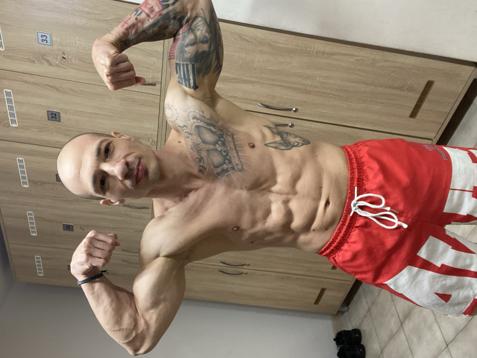 A shirtless man with a shaved head is flexing his biceps in what appears to be a locker room. He has numerous tattoos on his arms and chest. He is wearing red and white shorts with a drawstring. The locker room has light brown wooden lockers with silver handles, numbered 32 and 33. There is a pair of black shoes on the floor. The location is possibly a gym locker room in Pleven, Bulgaria, though this cannot be definitively determined from the image. The man's action is posing and showing off his physique.