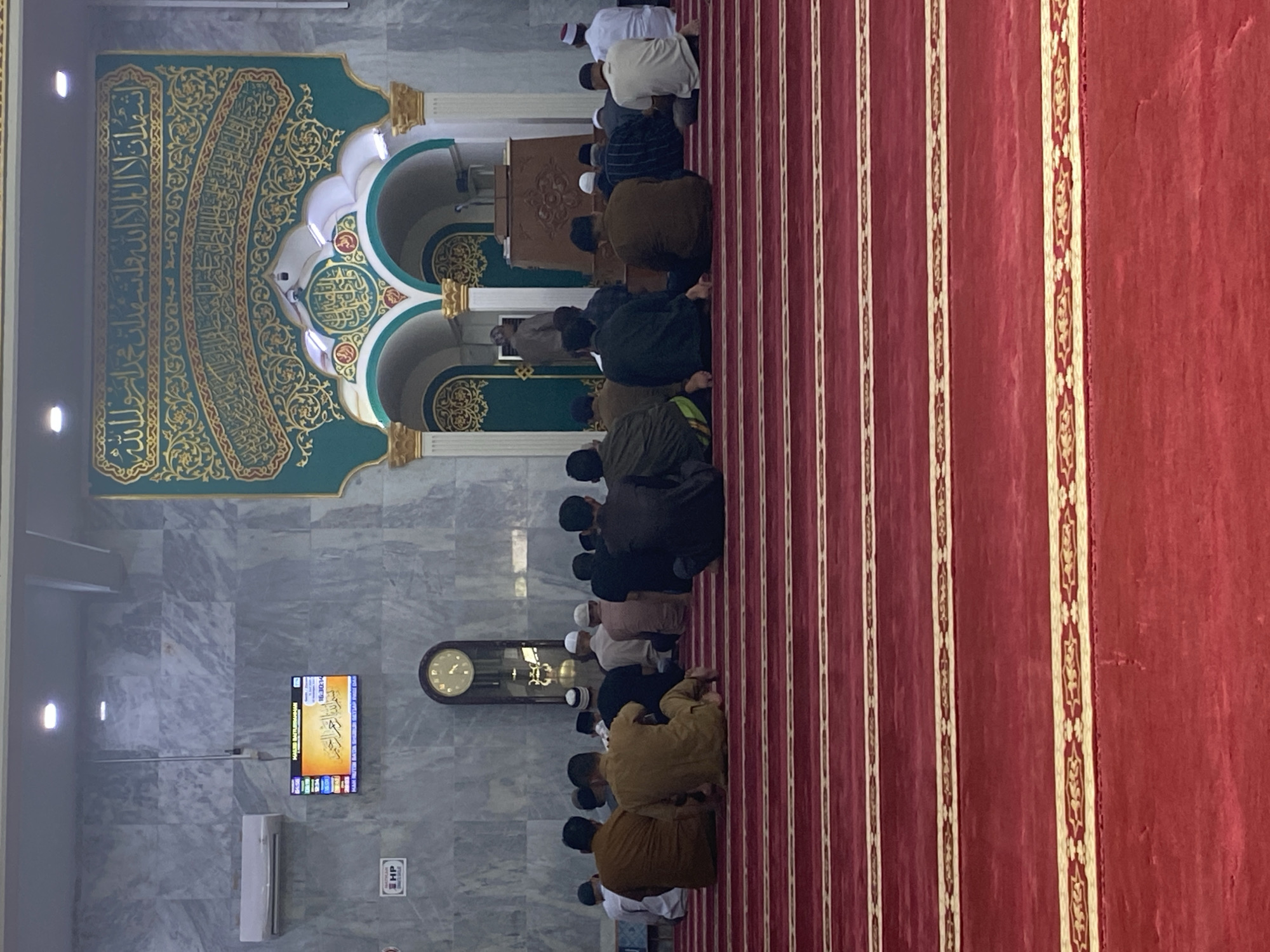 In Lhokseumawe, Indonesia, a group of men are seated on a red carpet in a mosque. The mosque features marble-patterned walls, a large clock, and ornate green and gold Islamic calligraphy above arched doorways. A digital display board, likely showing prayer times, is mounted on the wall. The men are facing towards the front of the mosque, with some appearing to be in prayer.
