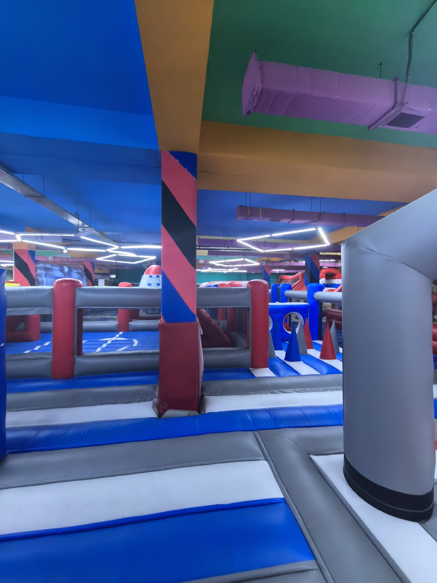 The image depicts the interior of an indoor inflatable playground. The environment is characterized by vibrant, primary colors, primarily blue, red, yellow, and green, adorning the ceiling and walls. Suspended from the ceiling are what appear to be air ducts covered in purple fabric, and modern linear lighting fixtures.
The playground itself is constructed from large, inflated structures. These structures are modular and interconnected, creating a multi-level play area with various elements such as slides, tunnels, and obstacle courses. The surfaces are predominantly made of durable, vinyl-like material in shades of blue, grey, and white, often arranged in striped or grid patterns. Red, cylindrical inflatable posts are visible throughout the play area, serving as structural supports or decorative elements. In the foreground, a large, grey inflatable archway dominates the right side of the frame, suggesting an entrance or a prominent feature of the playground. In the background, additional inflatable elements in red, white, and blue are visible, along with hints of other recreational structures and what might be a climbing wall.
The scene appears to be devoid of any people, suggesting it might be captured during off-hours, or perhaps it is a perspective that intentionally excludes visitors. The lighting is bright and even, typical of an indoor entertainment venue.
There is no information within the image to suggest a specific location in Fergana City, Uzbekistan. The visual elements are characteristic of a generic indoor inflatable play center found in many urban environments worldwide.