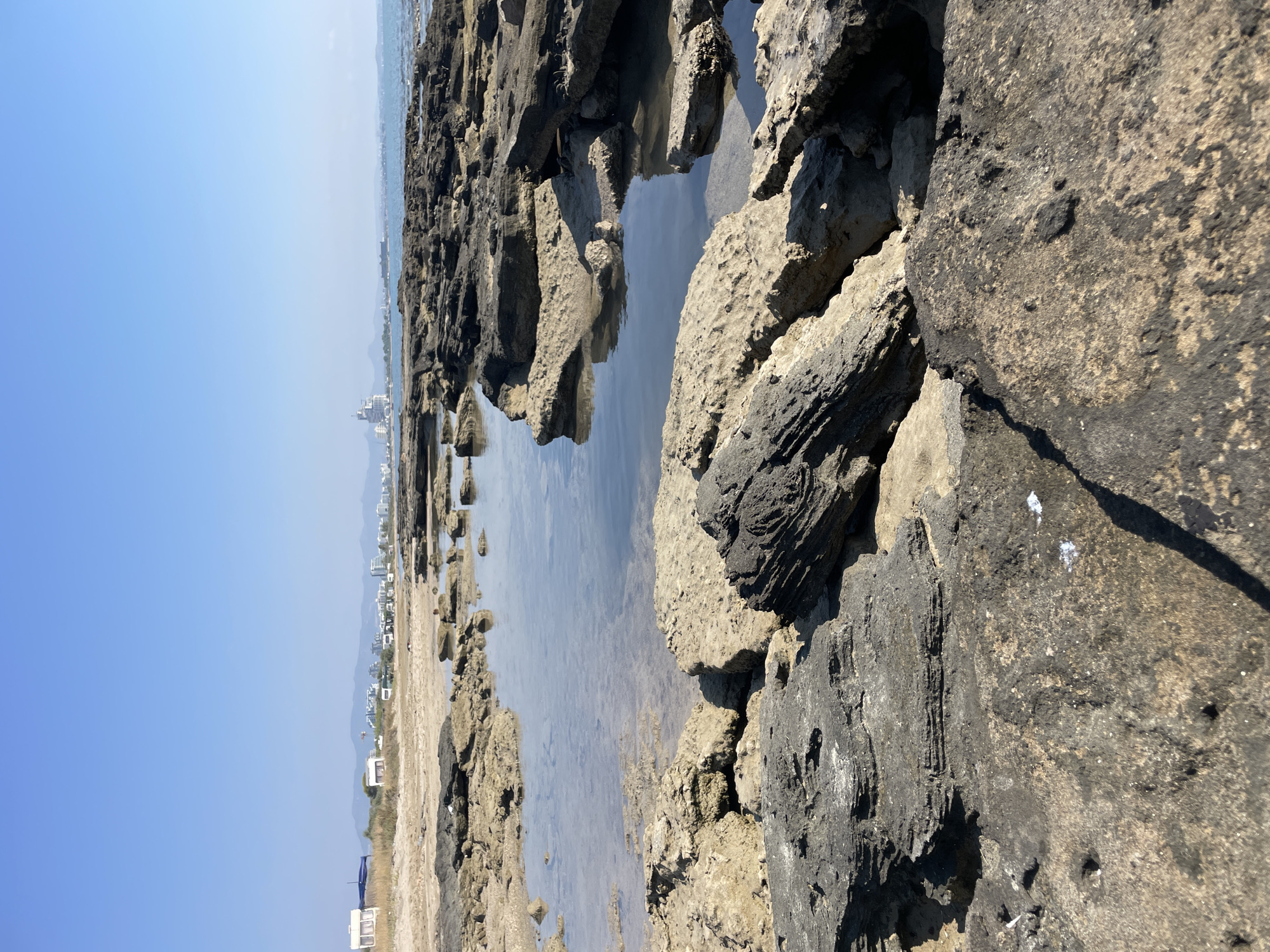 The image depicts a rocky coastline in Spathariko, Northern Cyprus, under a clear blue sky. In the foreground, large, dark, weathered rocks are interspersed with shallow pools of clear water reflecting the sky. The rocks show distinct layers and textures, with some appearing wet and others dry. Beyond the immediate rocky area, a sandy beach stretches back, dotted with scrubby vegetation. In the middle ground, several white caravans are visible on the beach, suggesting a campsite or holiday area. Further in the distance, a coastline with buildings, including a tall, modern white structure, lines the horizon. The sea is visible in the far background, calm and merging with the sky. The overall impression is a natural, somewhat rugged coastal landscape with signs of human presence.