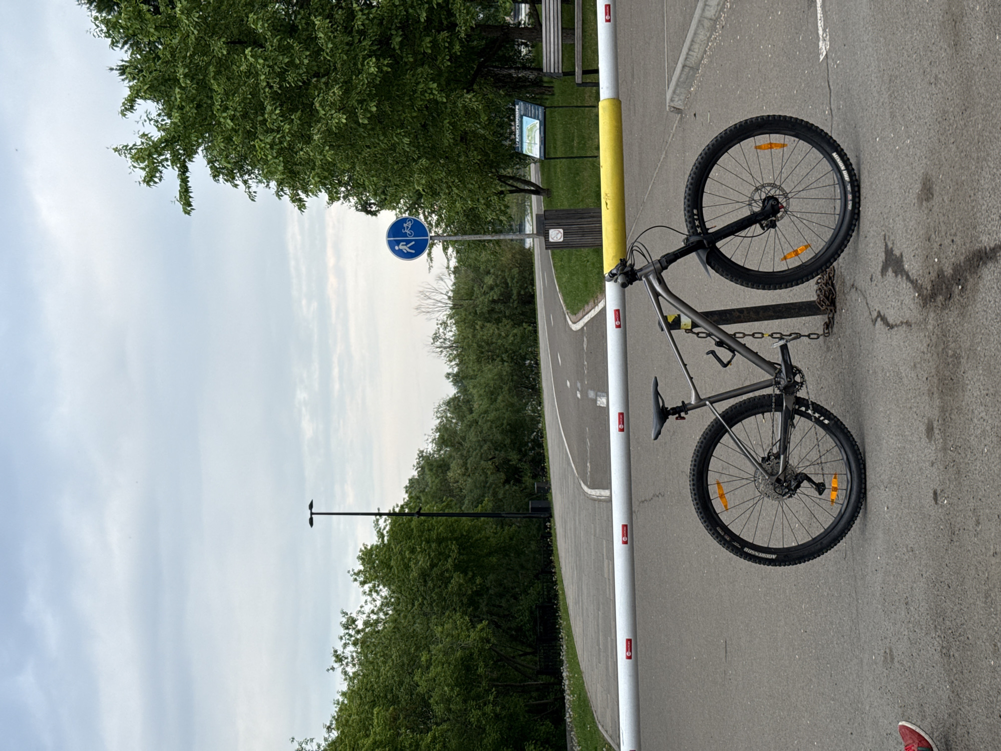 The image shows a mountain bike locked with a chain to a metal pole that is part of a retractable barrier across a paved path. The barrier is white with red circular logos and a yellow band. The path appears to be a dedicated bike and pedestrian route, indicated by a sign with blue pictograms of a person walking and a bicycle. This sign is on a pole to the right of the path. The path curves into the distance, bordered by trees and a cloudy sky. In the background, there is a trash can and a park information board. The location is identified as being in the city of Krasnoyarsk, Russia. No people are clearly visible, other than a red shoe in the bottom left corner, suggesting someone is just outside the frame. The scene appears to be in a park or recreational area.