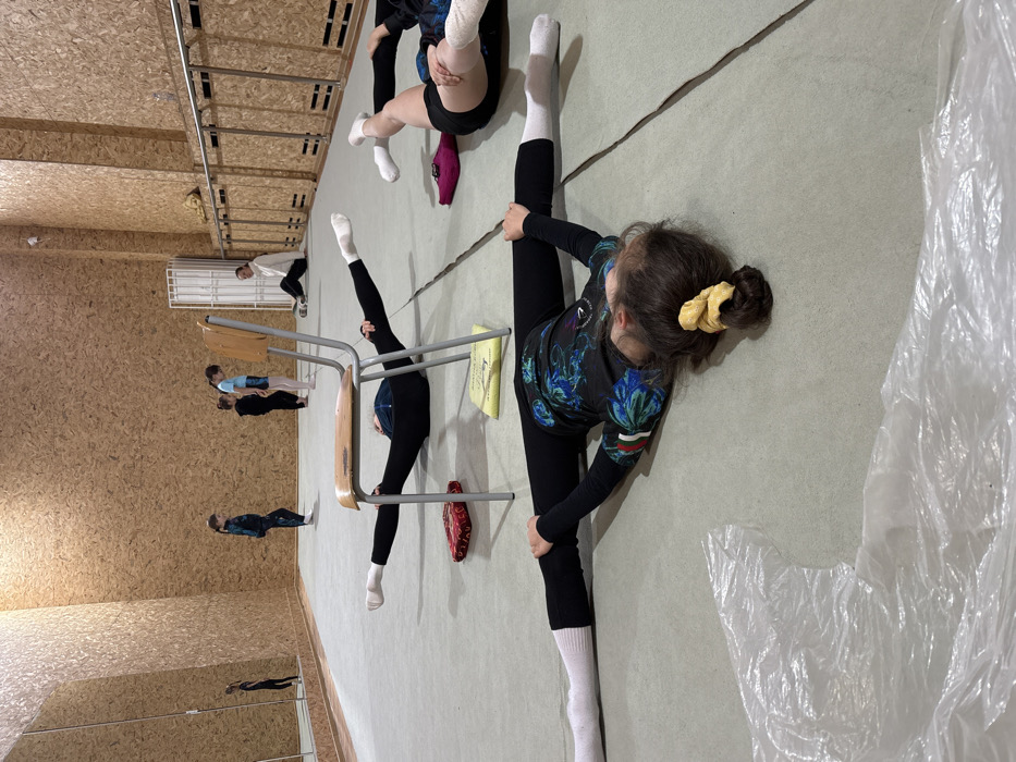 Gymnasts practice splits on a studio floor. One girl, wearing black leggings, a printed top, and a yellow scrunchie, performs a full split, touching the floor. Another gymnast uses a chair for support while doing a vertical split. A group of people watches nearby, reflected in a mirror along the wall. The studio has a wooden wall and a light-colored floor with visible cracks. Plastic covers a portion of the floor.