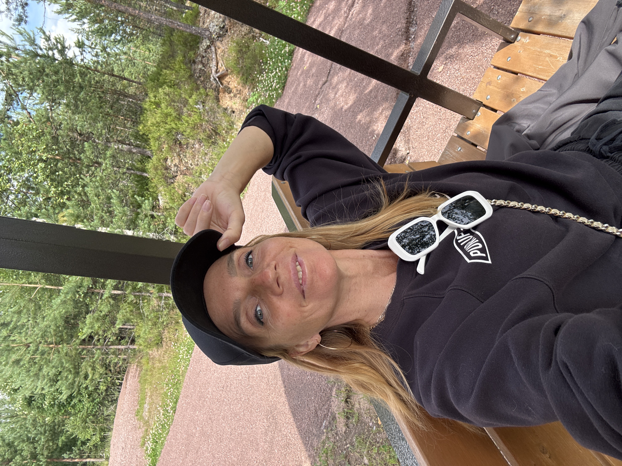 A blonde woman with fair skin and blue eyes wears a black baseball cap and a black sweatshirt. She has large hoop earrings and white sunglasses with dark lenses are hooked onto the collar of her sweatshirt. The woman is sitting on a wooden bench outdoors, with a wooded area and a reddish gravel path visible in the background. The woman is looking towards the camera with a slight smile and her right hand is resting on the brim of her cap. The location is identified as the city of Peski, Russia.
