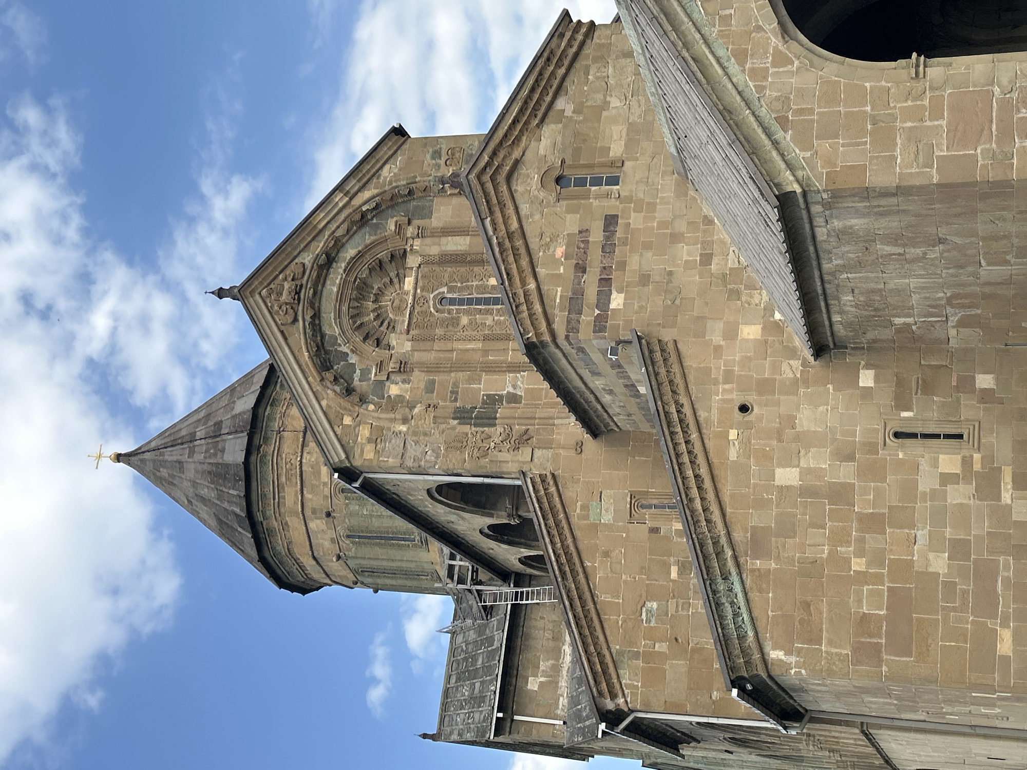 The image shows the Svetitskhoveli Cathedral, a Georgian Orthodox cathedral located in Mtskheta, Georgia. The cathedral is made of rough-hewn stone blocks and features a prominent conical dome topped with a golden cross. The facade is adorned with intricate carvings and arched windows. A metal ladder is propped against the side of the building, leaning towards the roof. The sky above is a bright blue with scattered white clouds.