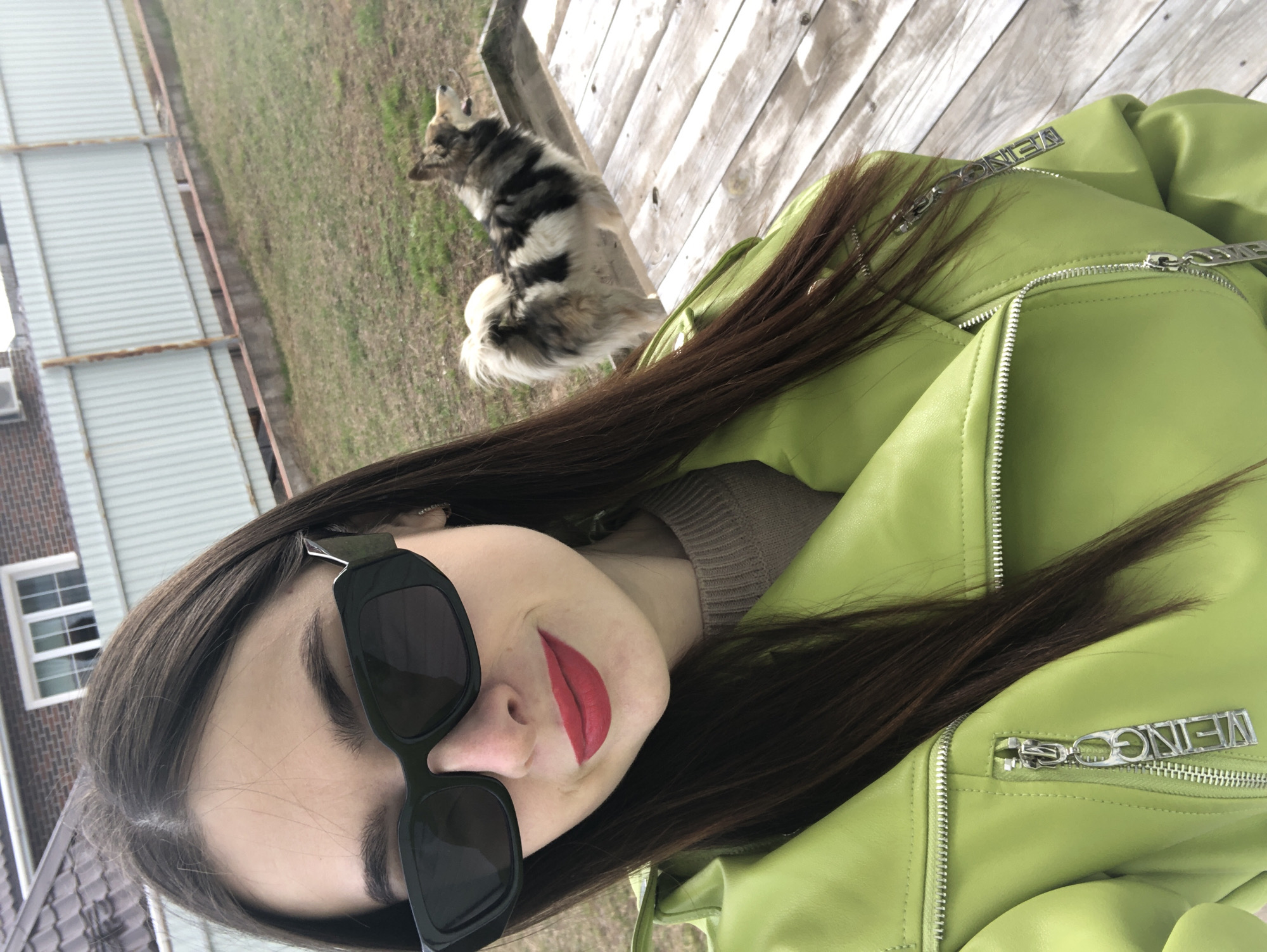 The image captures a tilted, close-up outdoor selfie of a young woman in Noviy, Russia. The primary subject is a woman from the chest up, featuring long, dark brown hair, bold red lipstick, and large black rectangular sunglasses. She is wearing a vibrant lime green (chartreuse) faux leather jacket with numerous visible silver zippers and a beige knit top underneath.
In the upper right background, a medium-sized dog with multi-colored fur (black, white, and brown/grey patches) is visible, lying on a light-colored wooden surface, possibly a deck or patio.
The setting appears to be a backyard or a semi-rural outdoor area during bright daylight, suggesting sunshine. The ground beyond the wooden surface is sparsely covered with dry, light brown grass, hinting at early spring or late autumn. To the left, a light grey corrugated metal fence or wall with rusty horizontal support beams is visible. Behind this, a part of a brown brick building with a white-framed window and a dark object (possibly an air conditioning unit) can be seen.
A notable detail is the word "WEING" embossed on a rectangular metal zipper pull on the woman's jacket, with parts of "MEING" or "WEING" also visible on another zipper pull. The overall impression is one of casual style captured in a personal, angled shot.