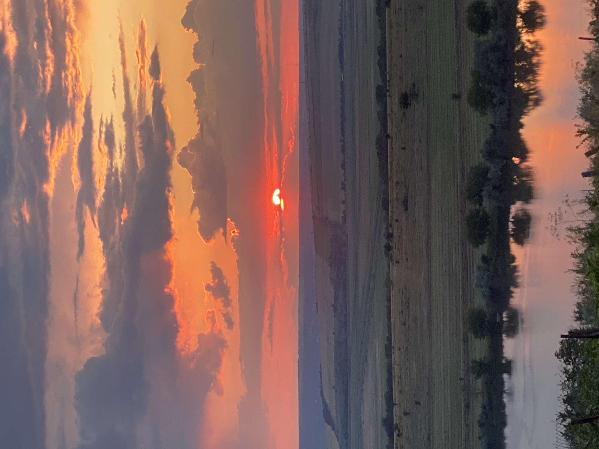 A serene sunset over a vast landscape in Cairaclia, Moldova. The sky is ablaze with vibrant orange and red hues, painted across a backdrop of dramatic clouds. The sun, a fiery orb, is partially obscured by a cloud bank, casting a warm glow on the scene. Below, rolling hills of green and brown stretch into the distance, dotted with scattered trees. A tranquil body of water in the foreground reflects the breathtaking colors of the sunset, creating a mirror image of the sky. The overall impression is one of peaceful natural beauty.