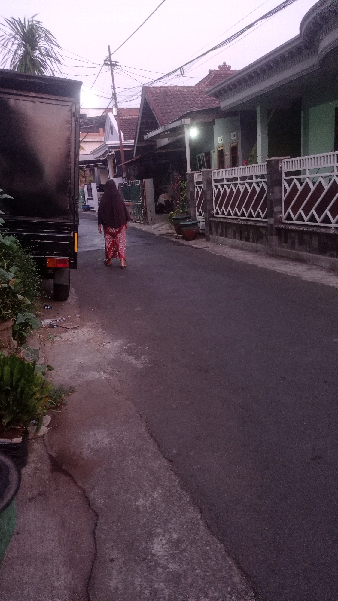 A woman wearing a dark headscarf and a patterned red and white skirt walks away from the camera down a residential street in Mulyoagung, Indonesia. She is walking on an asphalt road, with a concrete sidewalk on her left. Beside the sidewalk is a parked black truck, partially visible. The street is lined with houses, some with tiled roofs and white fences. The sky is overcast and the lighting suggests it is either early morning or late evening.