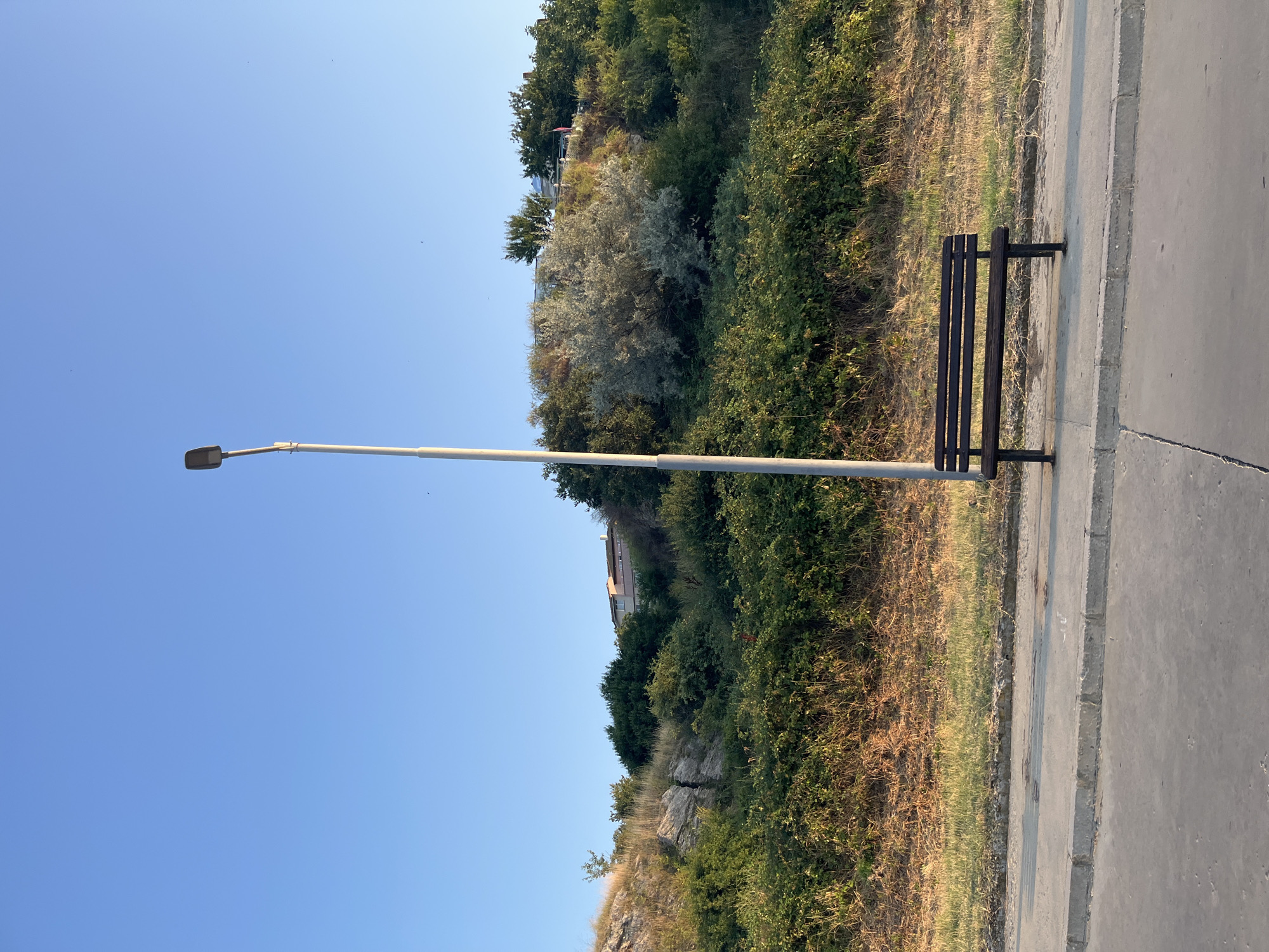 A solitary, dark wooden bench sits on a concrete sidewalk in Nesebar, Bulgaria. To the left of the bench, a tall, slender lamppost stands upright against a clear blue sky. Behind the sidewalk, a dry, grassy slope ascends to a dense green hillside covered with various types of foliage and shrubs. Further up the hill, a building with a pink roof is visible, nestled among the trees. The scene appears tranquil and bathed in natural light, suggesting it is daytime, likely late afternoon due to the gentle lighting.
