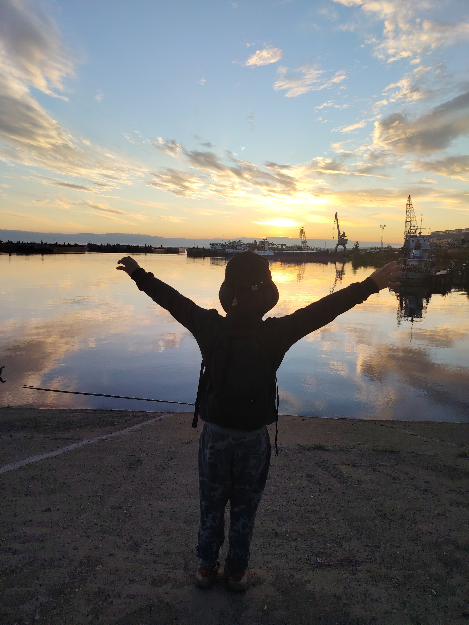 An individual stands on a concrete embankment, facing a body of water, with their arms outstretched towards a sunset sky. The sky is filled with scattered clouds, tinged with the colors of the setting sun. The water reflects the sky and the industrial landscape on the opposite bank, which features cranes and ships. The location is identifiable as a port area in the city of Chaikovsky, Russia, likely along the Kama River. The individual is silhouetted against the bright sky and is wearing a dark hooded jacket, camouflage pants, and a backpack. The ground in the foreground is rough and appears to be made of dirt and small stones.