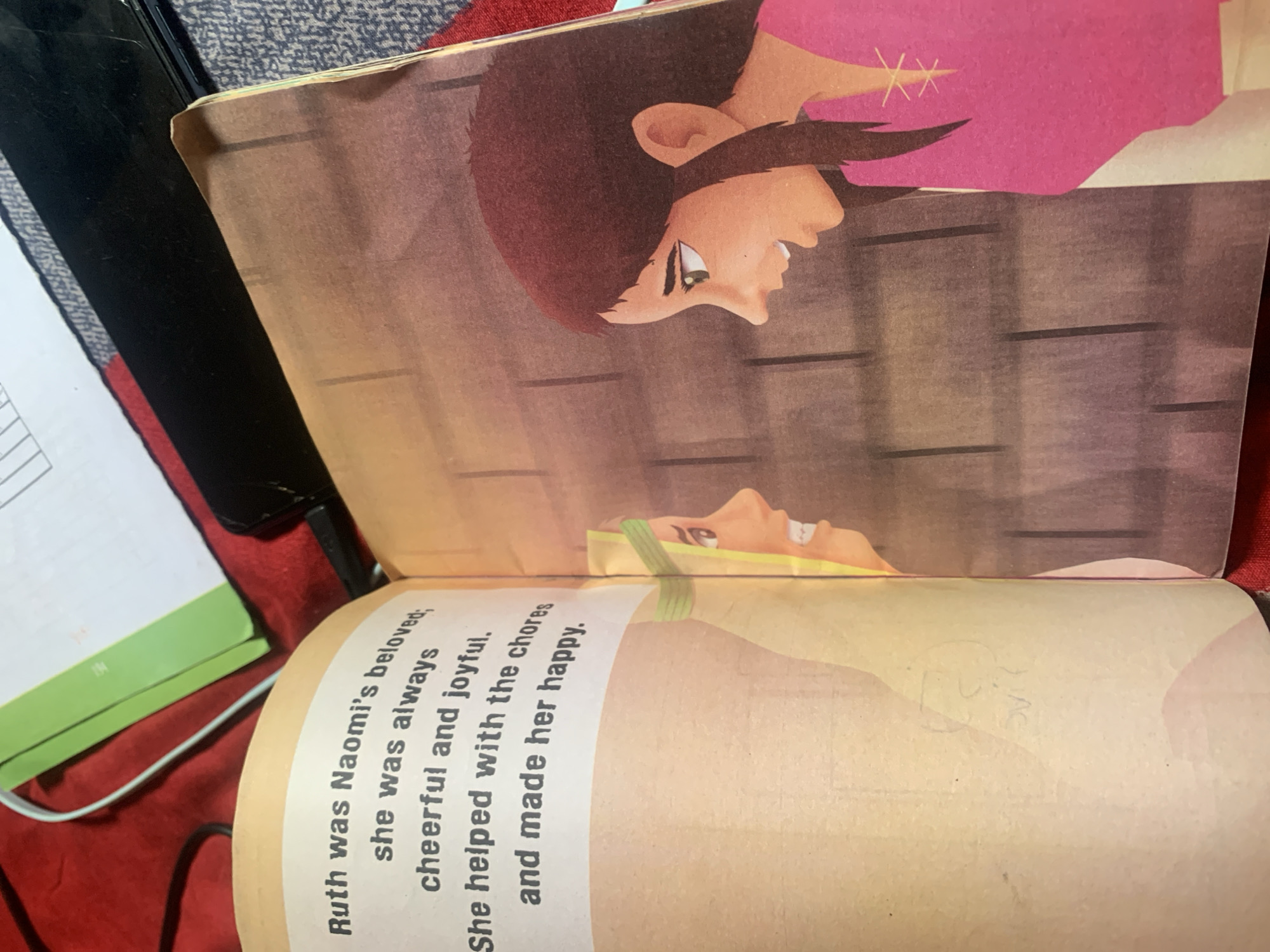 The image shows an open book with a story illustrated in a cartoon style. The top page of the book features an illustration of a person's face with dark hair, looking to the right. The person has an open mouth and appears to be smiling. They are wearing a pink garment. The background of this illustration appears to be a wooden wall. The bottom page of the book contains text that reads: "Ruth was Naomi's beloved: she was always cheerful and joyful, She helped with the chores and made her happy." Below this text, there is a faded drawing of a face. The book is lying on a red surface, possibly fabric, with some other objects, including a black object and a green object, visible in the background. The lighting suggests it is daytime, and the scene is indoors.