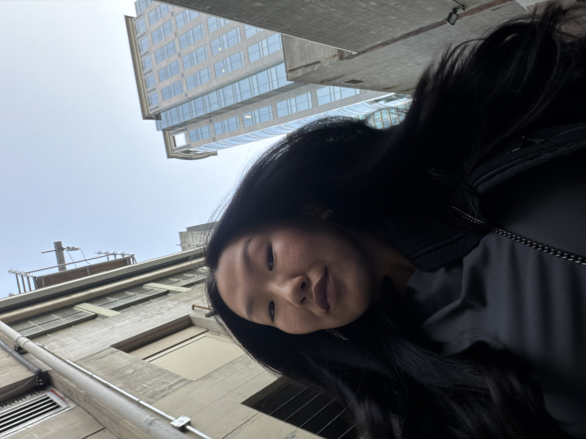 A low-angle, close-up selfie shows a woman with long, dark hair in an urban alleyway with tall buildings on either side. The woman's face is partially obscured by her hair and is looking up and slightly to her left. She is wearing a dark grey top and a black jacket. The background consists of concrete buildings, one of which is under construction with scaffolding. A modern glass skyscraper can be seen further in the distance. The scene is likely in Vancouver, Canada, given the architectural style and density of the buildings.