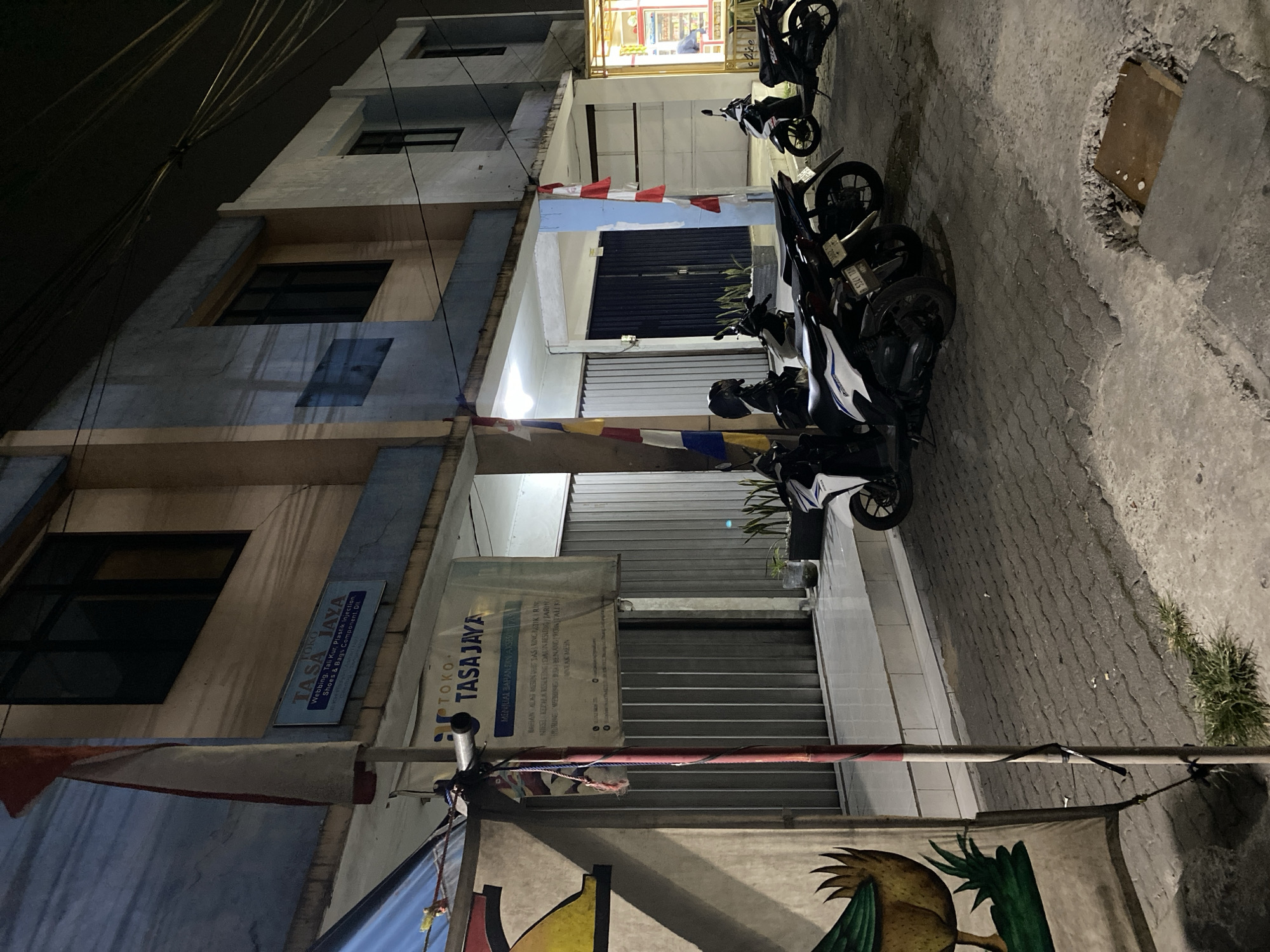 The image depicts the exterior of a building at night, identified as "TOKO TASA JAYA" located in Tangerang, Indonesia. The building appears to be a commercial establishment, with a blue and beige facade. Above the entrance, a sign reads "TOKO TASA JAYA" in blue text with a white background, along with smaller text detailing products like "Webbing, Tali Kur, Plastik Injection, Shoes & Bags Component, Oil". To the left of the sign, an Indonesian flag is partially visible, with its red and white colors distinct against the building's muted tones.
In front of the building, several motorcycles are parked on a paved walkway. The motorcycles are of various models and colors, including white, black, and blue. The environment suggests a street scene with overhead wires visible against the dark sky. The lighting is dim, characteristic of nighttime, with some artificial light illuminating the building's entrance and a portion of the street. A large banner with an illustration of a bird or chicken is also visible on the left side of the image, attached to a vertical pole. The overall scene is one of a quiet street in an Indonesian city at night, with commercial activity indicated by the shopfront and parked vehicles.