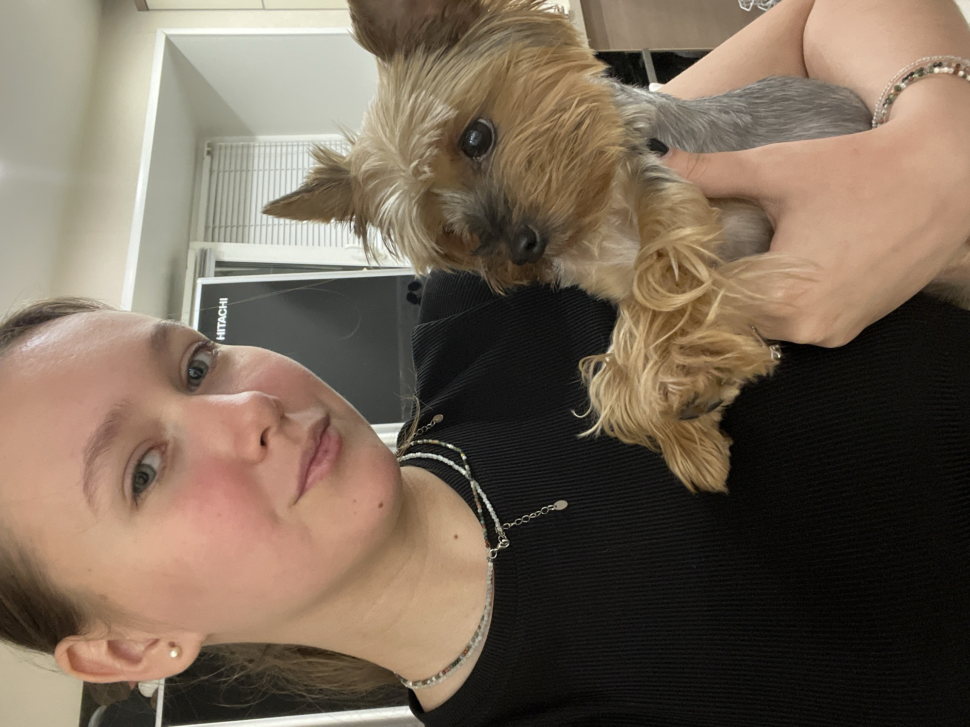 A selfie-style image shows a fair-skinned woman holding a Yorkshire Terrier-type dog. The woman is in the foreground, looking directly into the camera with a neutral expression. She has light eyes and fair hair, and wears a black top and delicate necklaces. Her skin appears slightly flushed.
The dog is held close to her chest, positioned between her face and her shoulder. It has brown and grey fur and is also looking at the camera. The woman's hand is visible, holding the dog.
The background is somewhat blurred, but it appears to be an indoor space. A black flat-screen monitor with a white "HITACHI" logo is behind the woman. There is also a white window or radiator cover, with a pale wall and cabinet doors above it.
There is no information available in the image about the location in the city of Chaikovsky, Russia.