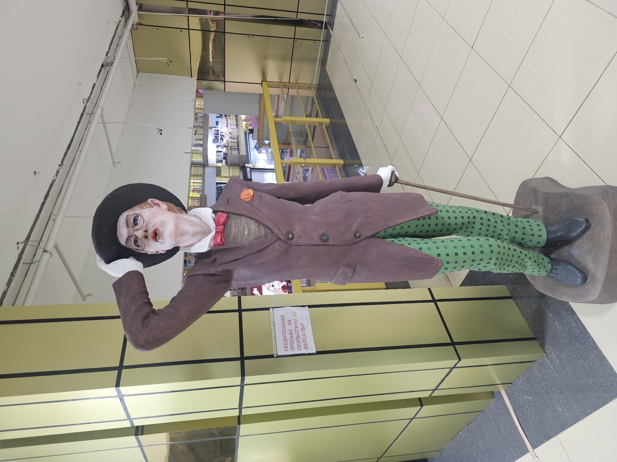 A full-body statue of a man dressed in a formal suit, a hat, and glasses stands on a gray base in an indoor environment. The statue is positioned near a pillar with a yellow and black striped design. A white sign with Russian text is attached to the pillar. The man statue wears a dark hat, glasses, a brown suit jacket with a red bow tie, and green checkered pants. He holds a cane in his right hand and has his left hand raised to his hat. In the background, a railing and several retail displays are visible, suggesting the location is a shopping mall. The floor is tiled with a light-colored pattern.