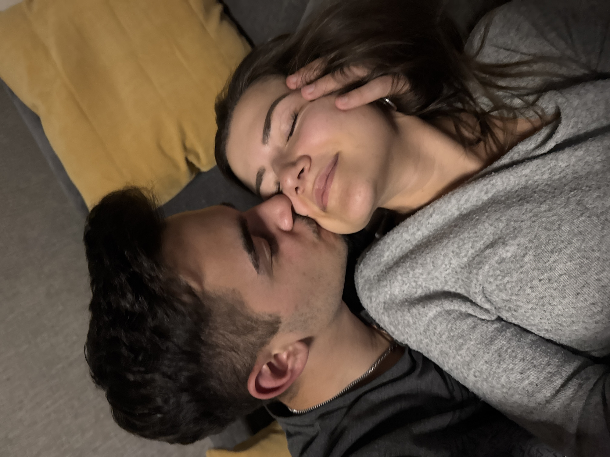 Two individuals, a man and a woman, are captured in a close, intimate embrace, appearing to be a couple. They are lying down together, likely on a couch or bed, against a backdrop of a soft yellow pillow on the left and grey fabric throughout.
The woman is positioned above or in front, with her head gently tilted, her eyes closed, and a soft, contented smile gracing her lips. Her dark hair cascades around her, and her right hand is lightly placed near her temple. She is wearing a light grey, textured sweater, and a silver ring is visible on her left ring finger.
The man is positioned beneath or behind her, with his face turned towards hers. He appears to be gently kissing or nuzzling her cheek or nose area. He has short dark hair and stubble, and is wearing a dark shirt with a silver chain around his neck. His eyes also appear to be closed or looking down, mirroring the woman's peaceful demeanor.
The scene conveys a moment of deep affection, comfort, and tenderness between the subjects. The lighting is dim and soft, suggesting an indoor setting during the evening or night, which enhances the intimate and cozy atmosphere. No visible text is present in the image.