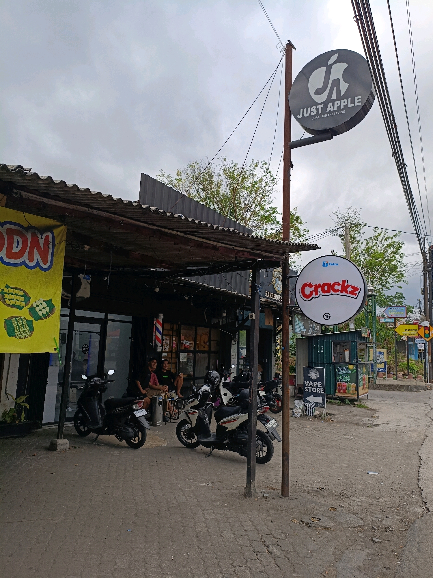 A wide outdoor shot captures a street scene in Ngaglik, Indonesia. The focus is on a row of shops, one of which has a corrugated metal roof. Two men are seated outside the shop, appearing to be engaged in conversation. In front of the shops, three motorcycles are parked.
Above the shops, two prominent signs are visible. The first, a circular sign with a dark gray background, displays the text "JUST APPLE" in white letters, along with a stylized apple logo. Below this, smaller text reads "JUAL - BELI - SERVICE". The second circular sign, also with a white background, features the word "Crackz" in red, bold lettering. A small "Tetra" logo is present at the top of this sign. A vertical pole supports both signs.
To the right of the shops, a vape store is indicated by a vertical sign. Further down the street, various other signs are visible, including directional signs and advertisements. The ground is paved with interlocking concrete blocks. The sky is overcast with gray clouds. A power line runs across the upper portion of the image.