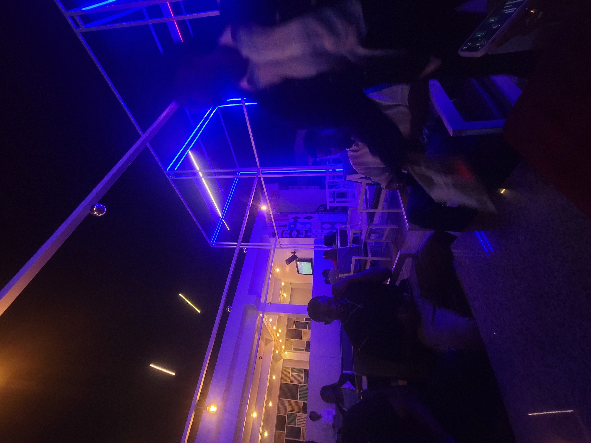 The image captures a nighttime scene at what appears to be an outdoor rooftop bar or lounge in Gura Top, Nigeria. The dominant colors are blue and purple, likely due to neon lighting decorating the structure. A partial view of a multi-story building is visible, illuminated with rows of vertical lights. People are present, with one individual close to the camera, partially in focus, dressed in dark clothing and a silver necklace. Other people are seen in the background near the building. A disco ball hangs from the rooftop structure. Tables and chairs are visible, indicating a social gathering or dining area. The overall scene suggests a vibrant, lively atmosphere.