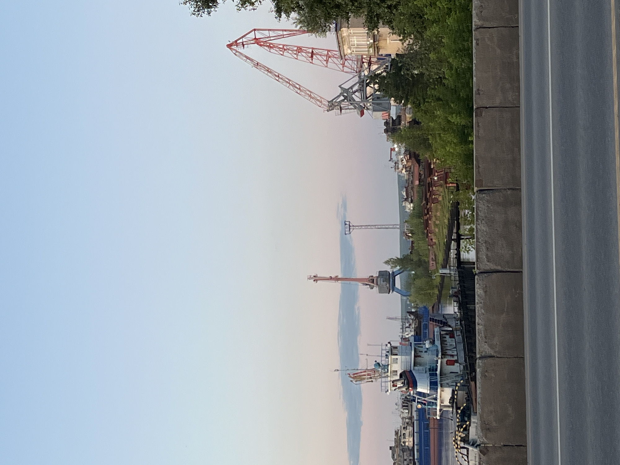 The image captures a wide view of a port area in Chaikovsky, Russia, during what appears to be late afternoon or early evening due to the soft, warm lighting. Dominating the scene are several large industrial cranes, some red and white, others blue, positioned among various vessels and structures. In the foreground, a concrete barrier separates a road from the port activities.
Several boats and barges are moored in the water. One prominent vessel in the mid-left is a white and blue tugboat with a detailed superstructure and several masts. Beyond this, a long blue barge is visible. Scattered throughout the port are numerous other ships, some appearing to be under repair or awaiting use, with rusty hulls and exposed metalwork.
The background shows a low skyline with trees and some buildings, including a light-colored structure with windows to the right of a prominent red crane. A tall, thin tower with lights, possibly a navigation or communication mast, stands in the distance. The sky is a gradient of pale blue and soft pink, indicating a peaceful and serene atmosphere. The overall impression is one of industrial activity in a maritime setting under a tranquil sky.