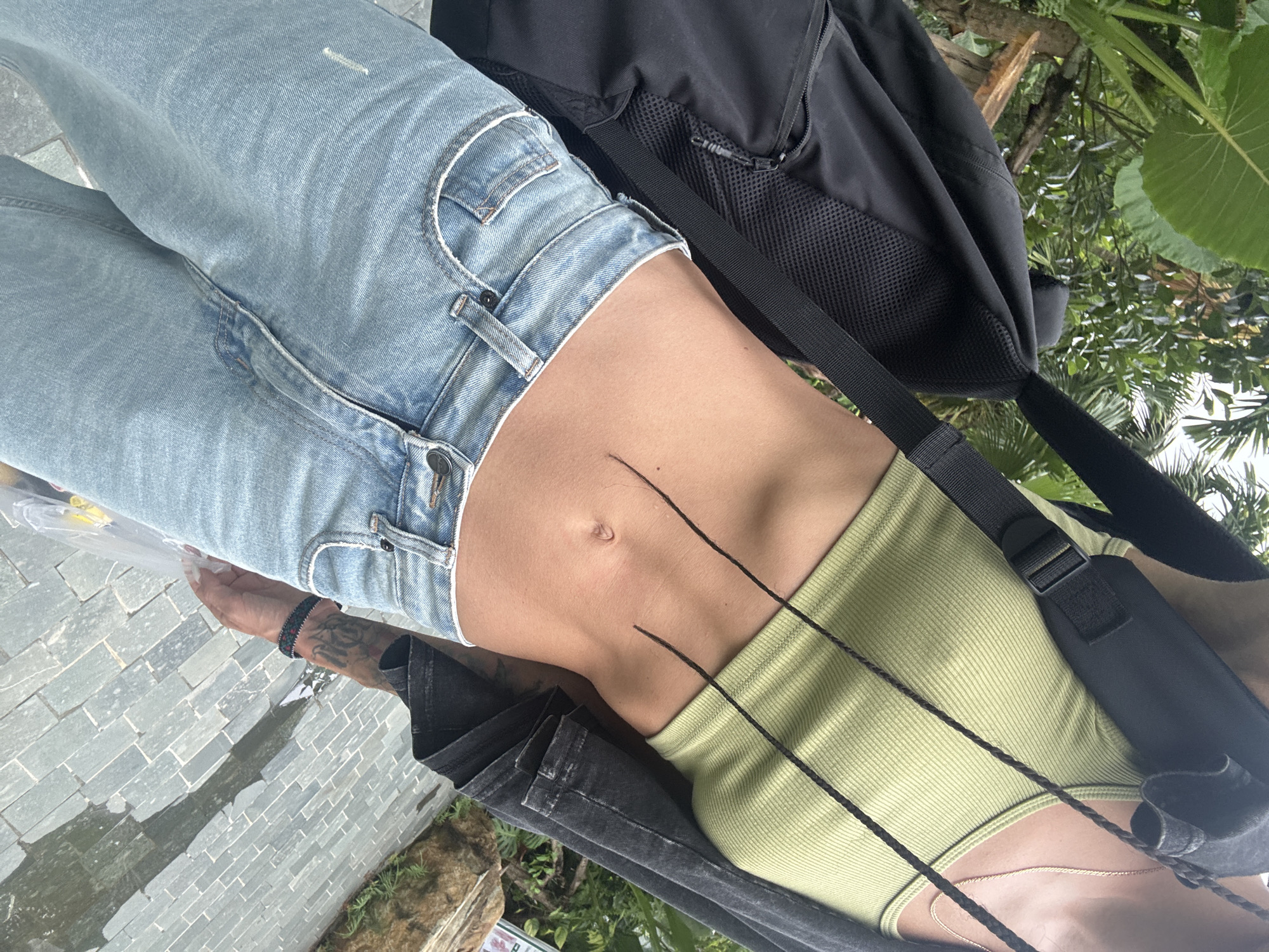 The image shows a person wearing light blue denim jeans and a lime green ribbed tank top. The person has a tattoo on their left arm and is wearing a bracelet. They are carrying a black backpack. The scene appears to be outdoors, with lush green foliage and large leaves visible in the background. The ground is paved with grey stone tiles, and there is evidence of water on the ground, suggesting it might have rained recently or there is a water feature nearby. The lighting suggests it is daytime, possibly with overcast skies due to the diffused light. The overall impression is one of a casual outing in a natural or semi-natural environment. There is no visible text in the image. The provided location context of Pù Luông Commune, Vietnam, suggests a tropical or subtropical setting.