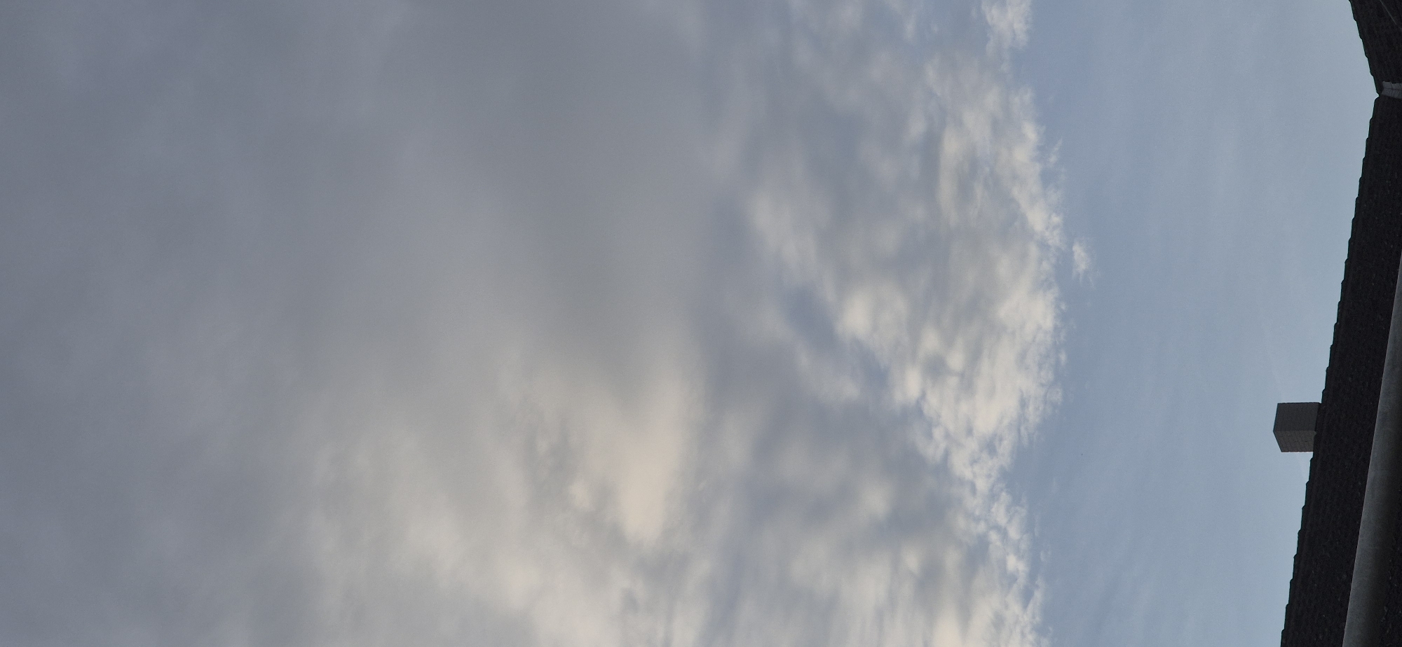The image shows a cloudy sky above the roof of a building with a chimney. The sky is a mix of gray and white clouds, with a clear blue sky visible in the lower portion. The roof is dark and appears to be made of shingles. The chimney is square and made of brick. The scene is likely a residential area in Mainz, Germany, given the architectural style of the roof and chimney. There are no people or animals visible in the image. The lighting suggests it might be early morning or late afternoon.