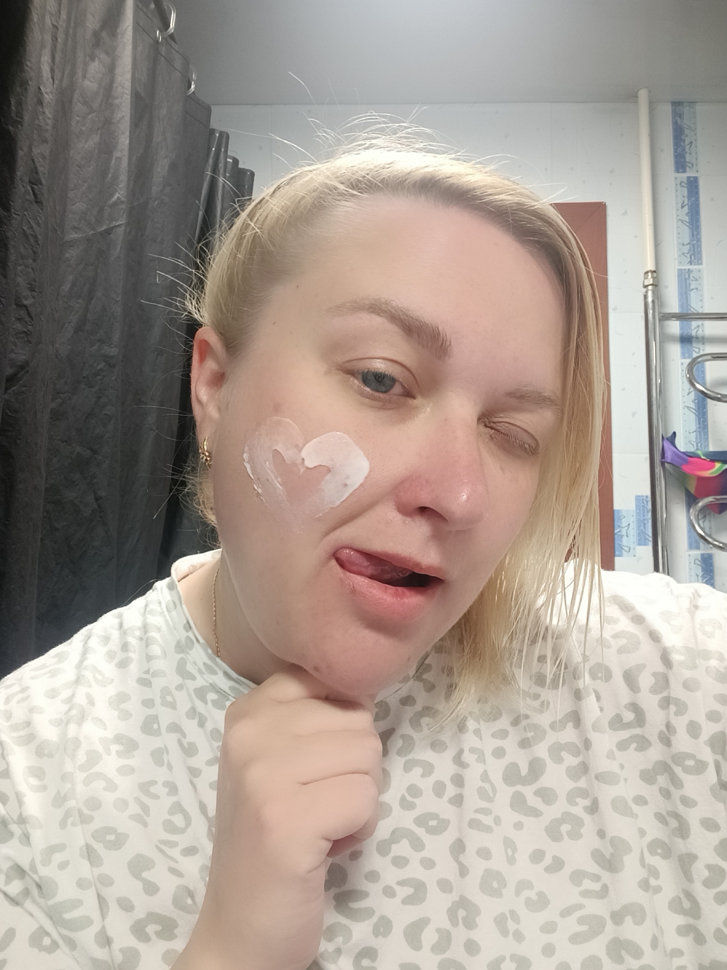 A woman with light blonde hair and blue eyes is posing for a selfie in what appears to be a bathroom. A heart-shaped blob of white cream is visible on her left cheek. Her left eye is partially closed, and her tongue is sticking out slightly. She is wearing a white and gray leopard-print top. Her left hand is raised near her chin.
A dark shower curtain is visible on the left side of the image. A white tiled wall with blue accents is visible on the right, along with a heated towel rack.
The location is the city of Chaikovsky, Russia.