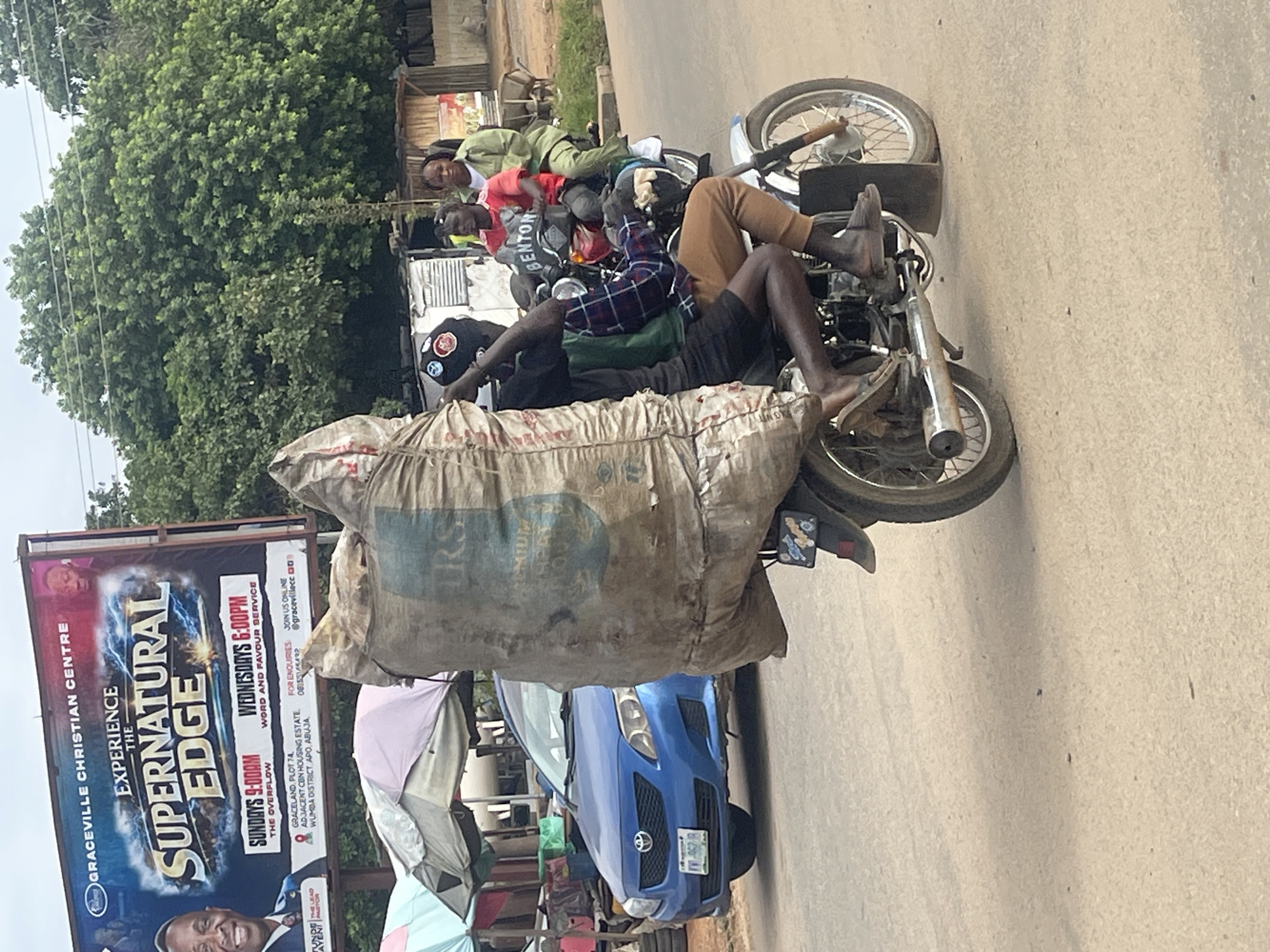 In Abuja, Nigeria, a motorcycle carries three people and a large sack. The driver, wearing a black cap and a plaid shirt over a green vest, steers the motorcycle. Behind him, a person in a red shirt with "BENTON" printed on it is seated, and another person in a green jacket is the rear passenger. The sack, made of rough material, is strapped to the back of the motorcycle and extends high above the riders. In the background, a blue Toyota Corolla is parked, and a large billboard for "GRACEVILLE CHRISTIAN CENTRE" advertises "EXPERIENCE THE SUPERNATURAL EDGE" with service times for Sundays and Wednesdays. Trees and various stalls are visible further in the background. The scene appears to be on a paved road with a dusty surface.