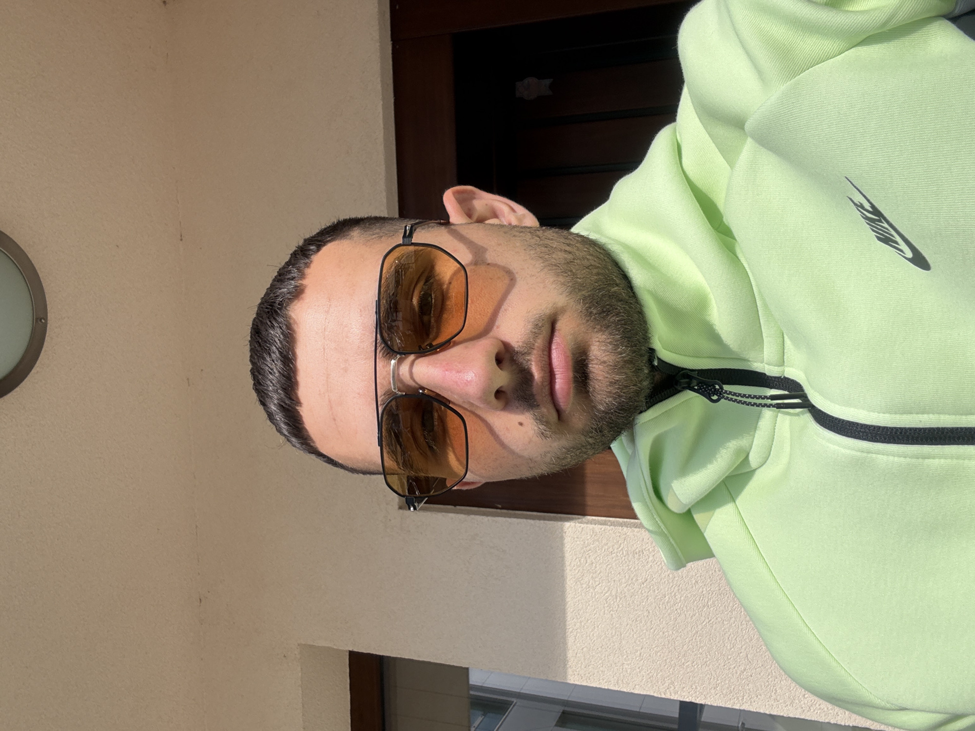 A man wearing a neon green Nike sweatshirt and brown tinted sunglasses is standing outside, likely on a balcony or porch. The background consists of a light beige wall, a window, and a dark-colored door. It is not possible to determine the location in the city of Gabrovo, Bulgaria from this image.