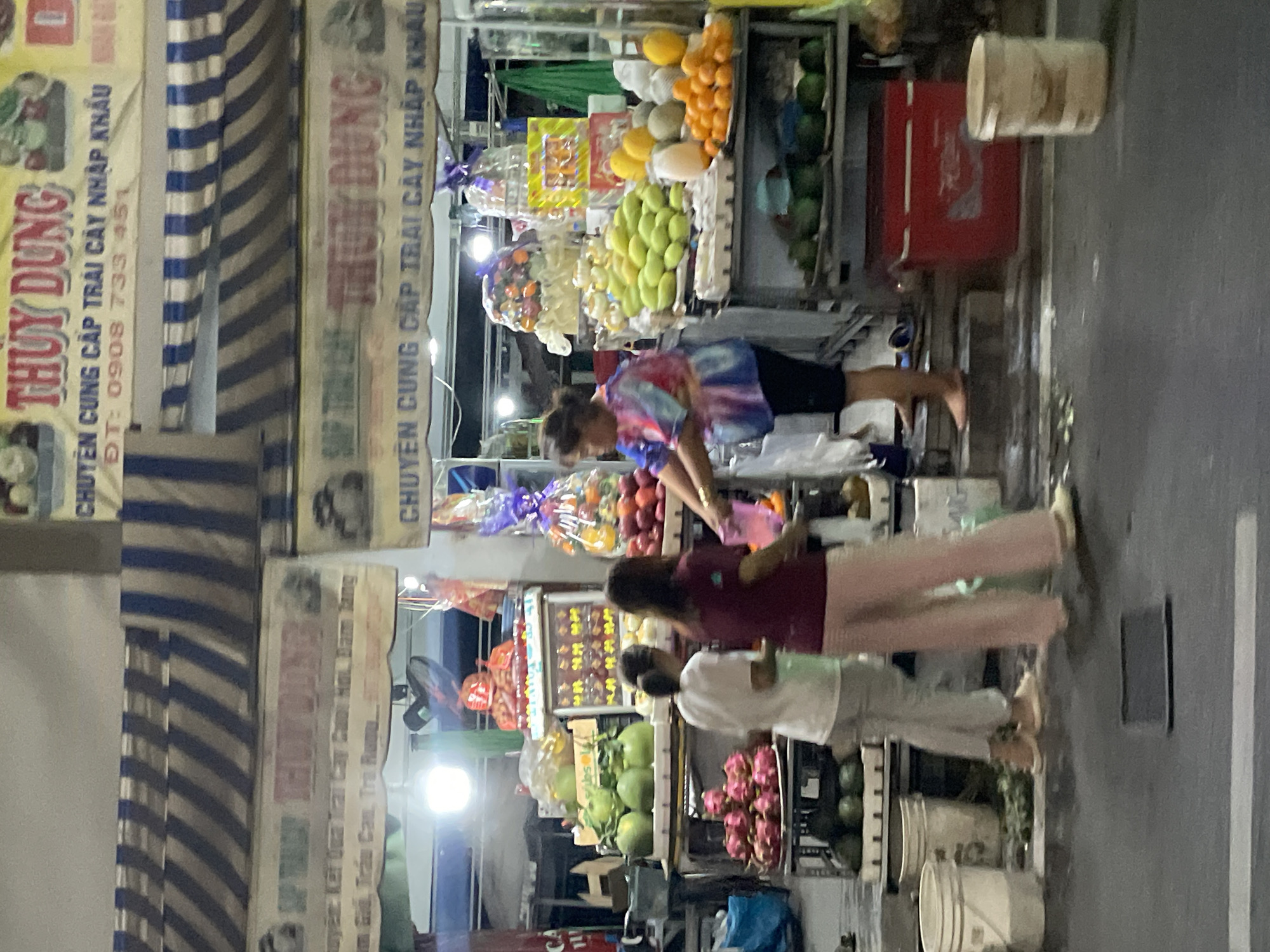The image depicts a fruit stand in Thủ Đức City, Vietnam. Two women are seen browsing the fruits, with one woman in a maroon shirt and pink pants interacting with another woman who is out of frame to the right. The fruit stand is well-stocked with various fruits, including dragon fruit, mangoes, oranges, and pomelos, arranged on shelves and in baskets. A large red cooler is visible behind the women. The scene appears to be outdoors, with a striped awning overhead providing shade. The ground in front of the stand is wet, possibly from rain or cleaning. The overall impression is a bustling market scene at dusk.