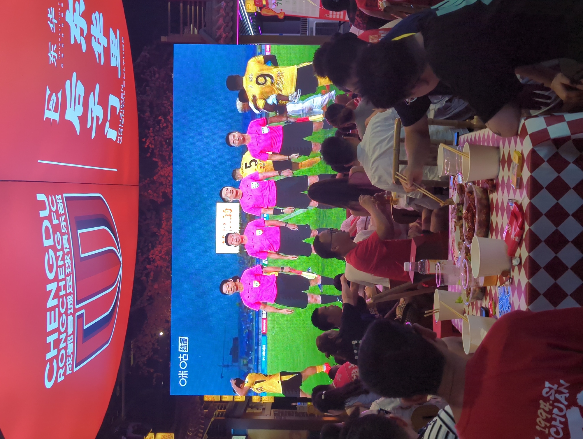 In the Qingyang District of China, a large screen displays a football match, showing referees in pink jerseys and players in yellow. The screen is mounted on a red banner with white text, indicating "CHENGDU RONGCHENG FC" and Chinese characters. Below the screen, a crowd of people is seated at tables covered with red and white checkered tablecloths, eating and watching the game. Some individuals are holding chopsticks, interacting with food, or looking at the screen. The environment appears to be an outdoor dining or viewing area, possibly a restaurant or street food stall, with traditional Chinese architectural elements visible in the background. The lighting suggests it is evening or nighttime.