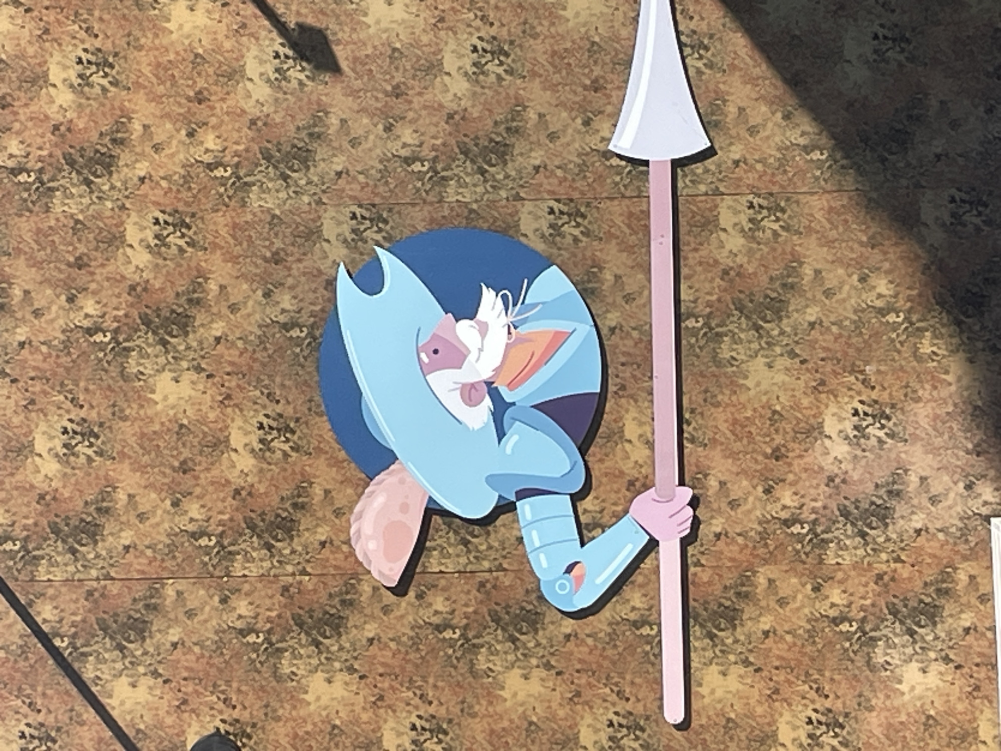 The image displays a stylized, cartoonish cutout or decal of a figure resembling a medieval knight or guard, possibly inspired by Don Quixote. The character is depicted in light blue armor with a prominent white beard or hair, a large pointed nose, and a distinct pink/peach-colored ear. It wears a light blue helmet with an exaggerated, pointed crest. The figure holds a long, slender spear or lance, which is light pink with a white, triangular head, held firmly in its gauntleted hand.
The background features a mottled, textured pattern in shades of brown, orange, and black, resembling a stone wall or patterned surface. The scene is lit by strong, directional lighting, casting distinct dark shadows on the background, including a large, angled shadow in the upper right and a more subtle shadow behind the figure itself. This indicates an outdoor setting during the daytime. The figure is in a static pose, not engaged in any active event. The location context for this image is Sochi, Russia. There is no visible text within the image.