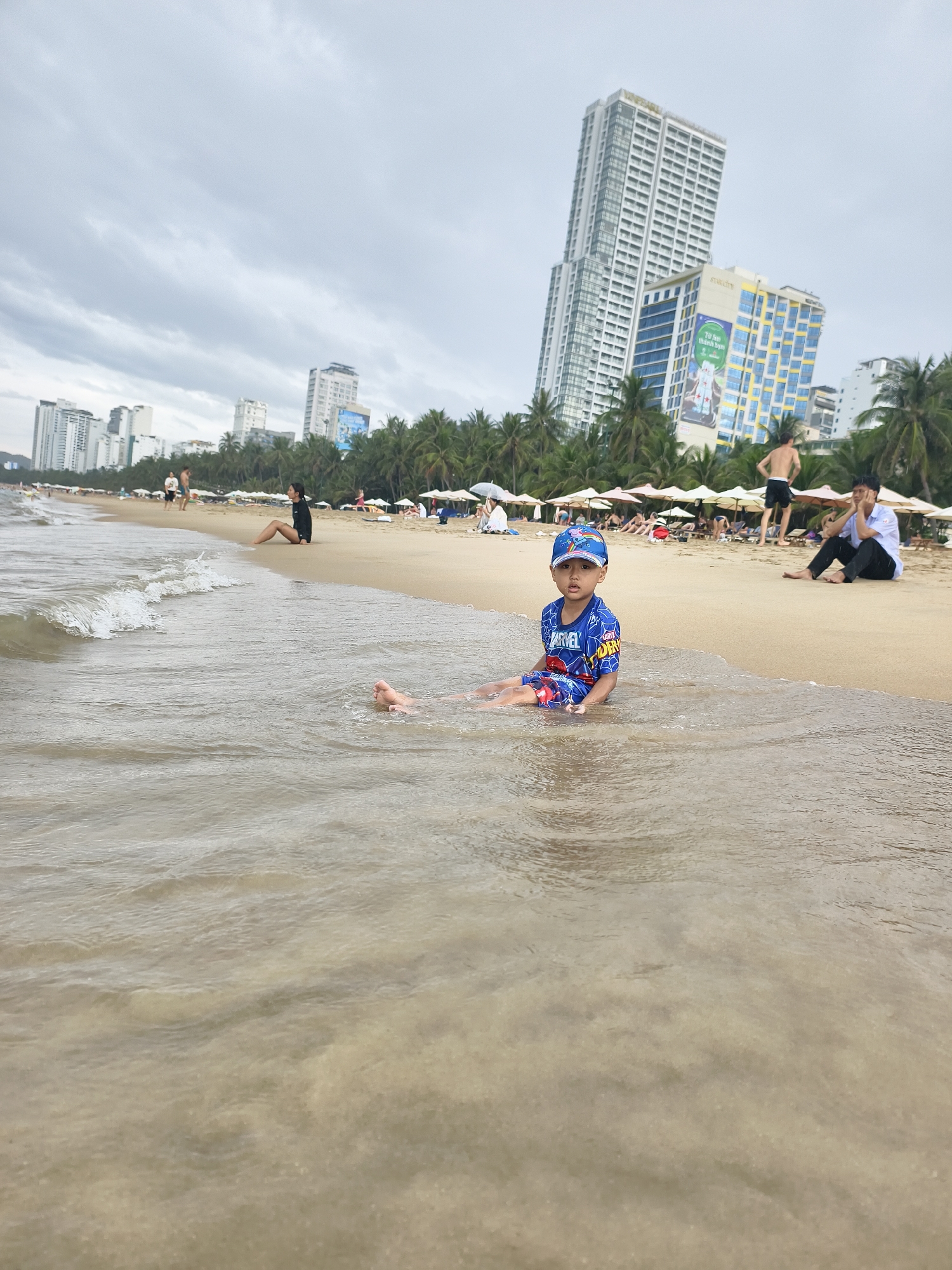 This image depicts a vibrant city beach scene on an overcast day. In the immediate foreground, a young boy is seated in the shallow, sandy-bottomed ocean water, looking directly at the camera with a neutral expression. He is wearing a blue swimwear set, including a long-sleeved top and cap, both featuring "MARVEL" and "SPIDER-MAN" branding.
Behind the child, the beach stretches back with gentle waves lapping the shore. Several adults are visible on the sand: one woman in a black long-sleeved swimsuit sits on the left, facing away from the camera, while further right, a man in black shorts stands, and another man in a white shirt and black trousers sits, possibly looking at a phone. In the background, numerous beach umbrellas and lounge chairs line the sandy shore, indicating a populated recreational area.
The setting is characterized by a dense line of palm trees that separates the beach from an urban landscape. Beyond the palm trees, a skyline of tall, modern high-rise buildings and skyscrapers is prominent, suggesting a bustling coastal city. The sky is uniformly covered with light gray clouds, indicating an overcast or cloudy day. A large billboard on one of the buildings clearly displays the text "STAR CITY" and "Tivi free, Không tiền điện" (meaning "Free TV, No electricity bill" in Vietnamese), serving as a strong location cue for Vietnam. The scene captures a moment of leisure at a developed beach resort.