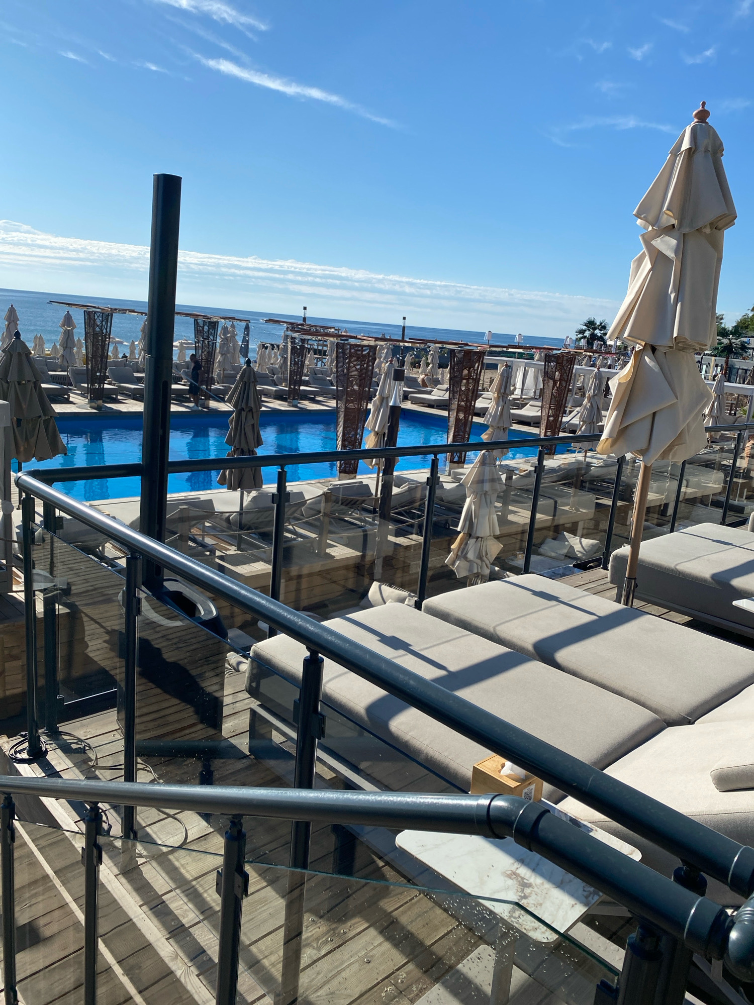 This is an outdoor photograph taken on a sunny day, showing a resort-style swimming pool area. The pool is filled with bright blue water and is surrounded by numerous lounge chairs and umbrellas, many of which are closed. In the foreground, there is a wooden deck with a railing made of metal and glass. Several plush lounge chairs are arranged on the deck, some of which are occupied. The railing and deck suggest an elevated viewing area overlooking the pool and the sea beyond. The ocean is visible in the background, meeting the clear blue sky. The overall scene depicts a leisure environment, likely a beach club or hotel pool.