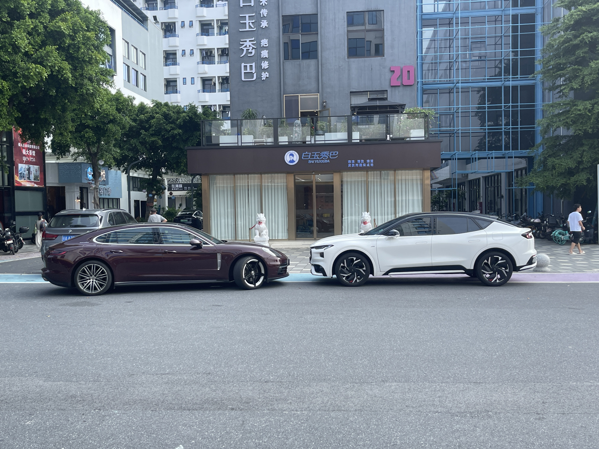 In Panyu District, China, on a city street, two cars are parked side-by-side. The car on the left is a dark red Porsche Panamera, and the car on the right is a white electric vehicle. The street is paved with asphalt and has a white and purple striped pedestrian crossing. In the background, there are multi-story buildings, including a building with a blue glass facade and another building with a gray facade featuring Chinese characters and a pink "20." Trees line the street, and there are some parked motorcycles and bicycles. Two people in white suits stand near the entrance of a building with the text "BAI YU XIU BA" and a circular logo with a profile of a man. Another person is walking on the sidewalk in the background.
