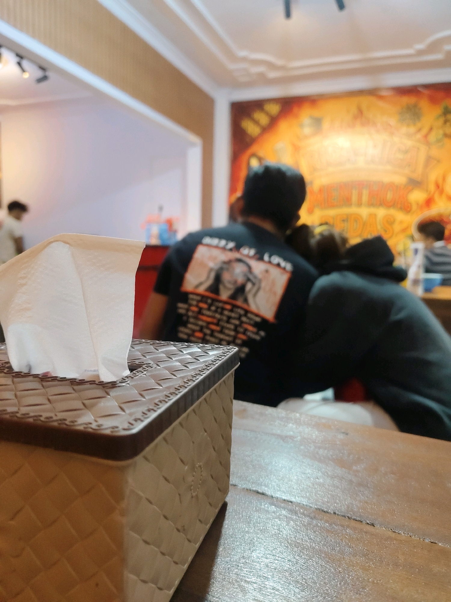 A close-up shot from a low angle focuses on a tissue box with a white tissue emerging from it. The tissue box is positioned on a wooden table. In the background, out of focus, two individuals are seen. One person, wearing a black t-shirt with a graphic print, is seated, with their head resting on the shoulder of another person who is wearing a dark hooded jacket and appears to be embracing them. Behind them, a colorful mural is visible on the wall, depicting text that seems to be in Indonesian, including "RICA-RICA" and "MENTHOK PEDAS". To the left, another blurry figure is partially visible, and further back, part of a white wall and some ceiling lights can be seen. The overall setting suggests an indoor dining or casual establishment, potentially a warung or restaurant in Probolinggo, Indonesia, known for its local cuisine.