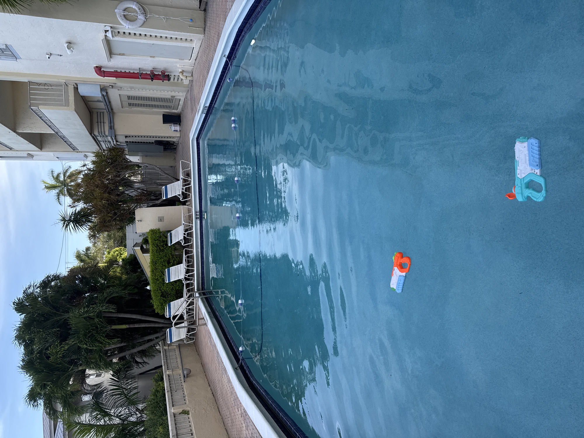 The image depicts an outdoor swimming pool at a residential building in Fort Lauderdale, Florida. The pool has clear blue water and a tiled edge. There are lounge chairs arranged around the pool, and palm trees and a building are visible in the background. Two water guns are floating in the pool.