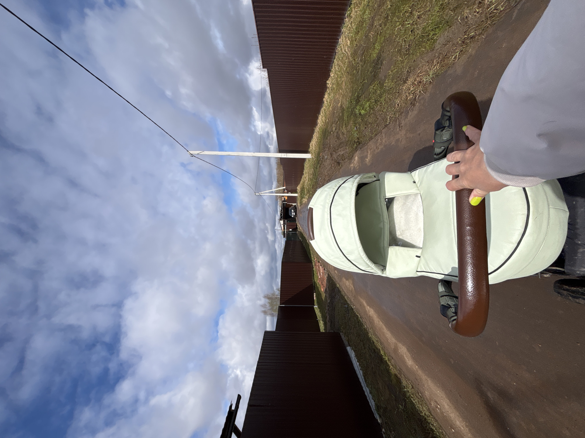 A person is pushing a light green stroller with a brown handle down a dirt road in Раздолье, Russia. The road is flanked by a tall, dark brown fence on either side. To the left, utility poles and electrical lines are visible against a sky filled with white and gray clouds. A building is seen in the distance at the end of the road. The ground is partly covered with grass and mud. A hand is on the stroller handle.