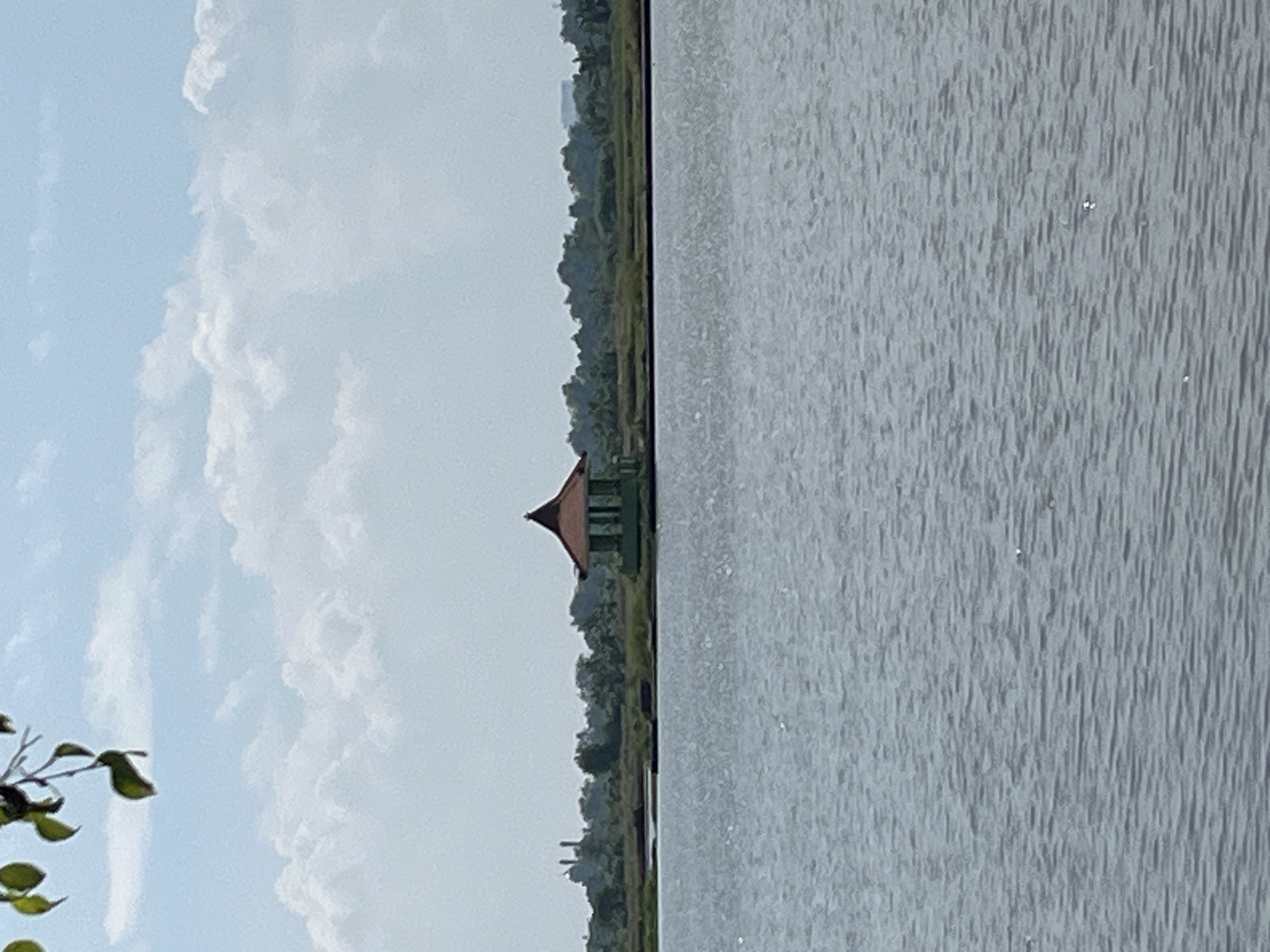 A tranquil body of water occupies the foreground of the image, its surface rippled by a gentle breeze. In the middle ground, a small, octagonal gazebo with a reddish-brown, tiered roof stands on a patch of grassy land. The gazebo has green, vertical posts and appears to be open on all sides. Beyond the gazebo, a line of lush green trees forms the backdrop, with some distant structures, possibly industrial, visible to the left. The sky above is a pale blue, dotted with fluffy white clouds. In the upper left corner, a branch with green leaves is partially visible, framing the scene. The overall impression is one of a peaceful, natural setting within a developed area, likely located in the city of Pesawahan, Indonesia, given the characteristic tropical flora and building styles.