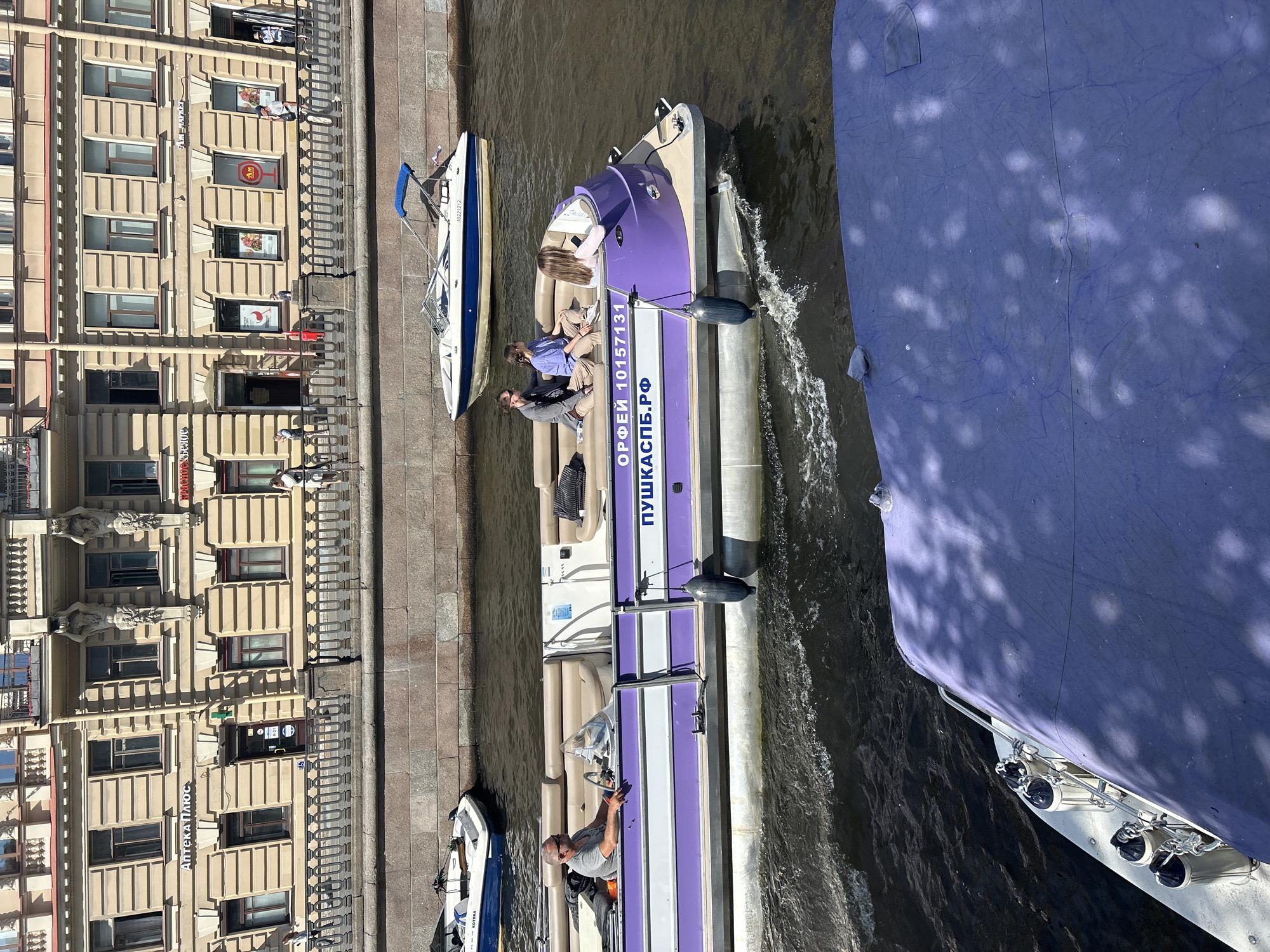 In an outdoor urban canal scene, a purple and white pontoon boat moves through dark, rippling water. The boat has a light-colored interior with several people onboard. A man wearing sunglasses and a gray t-shirt is in the driver's seat. Two women are seated behind him. The pontoon boat's hull is marked with Cyrillic text, "ОРФЕЙ 10157131" and "ШКАСПБ.РФ". Two black fenders hang from the side of the boat. In the background, a tan stone embankment lines the canal, topped with a decorative metal railing. Behind the embankment, a multi-story building with many windows and architectural detailing stands. Another white motorboat with a blue canopy is docked further down the canal, and a portion of a blue-covered boat is visible in the foreground. The scene is illuminated by bright sunlight, casting shadows on the water and the boat in the foreground.