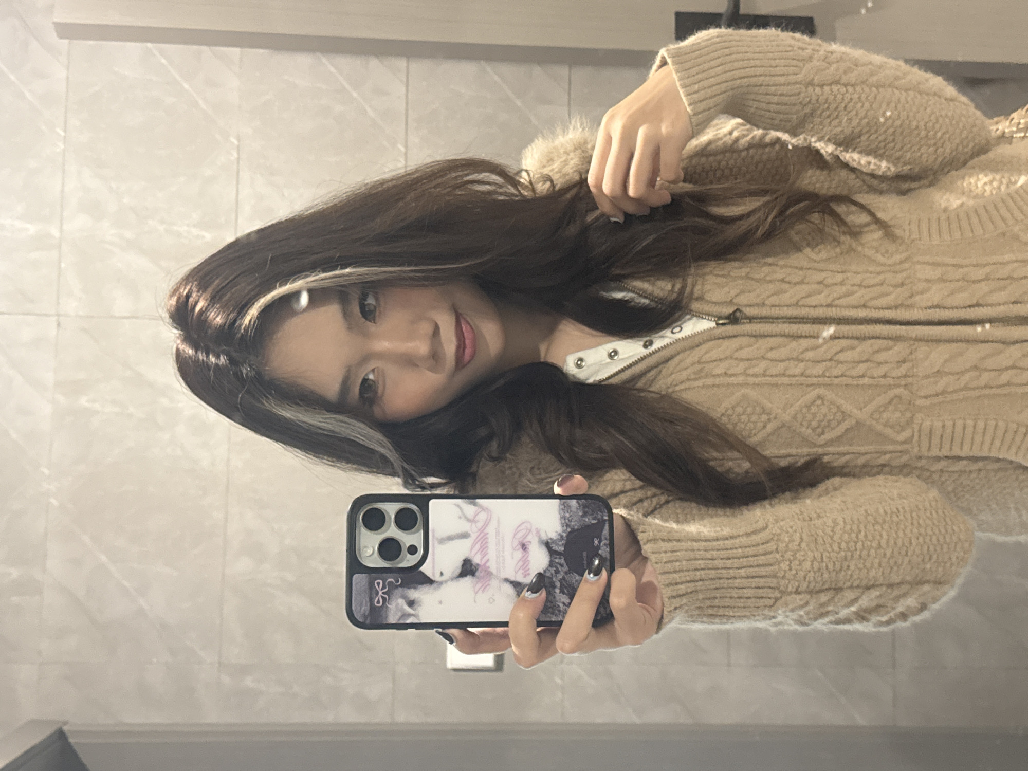 An East Asian female is depicted from the chest up, reflected in a mirror within an indoor setting in Nan'an District, China. She holds a dark-colored smartphone, likely an iPhone with a three-lens camera array, in her left hand (viewer's right) to capture her reflection. The phone is fitted with a case featuring a monochrome image, light pink text, and a small pink bow graphic. Her right hand (viewer's left) is positioned near her shoulder, lightly touching her long, wavy, dark brown hair which has prominent lighter streaks framing her face. She is wearing a light beige or tan cable-knit cardigan with a fuzzy collar, partially unzipped to reveal a white, buttoned or snapped shirt underneath. Her nails are painted dark with metallic tips. She exhibits a slight, closed-lip smile and looks directly towards the phone/camera. The background reflection consists of light-colored, possibly white or light gray, rectangular wall tiles with a subtle veining pattern. A dark vertical element and a dark door handle are visible on the far right of the reflection.