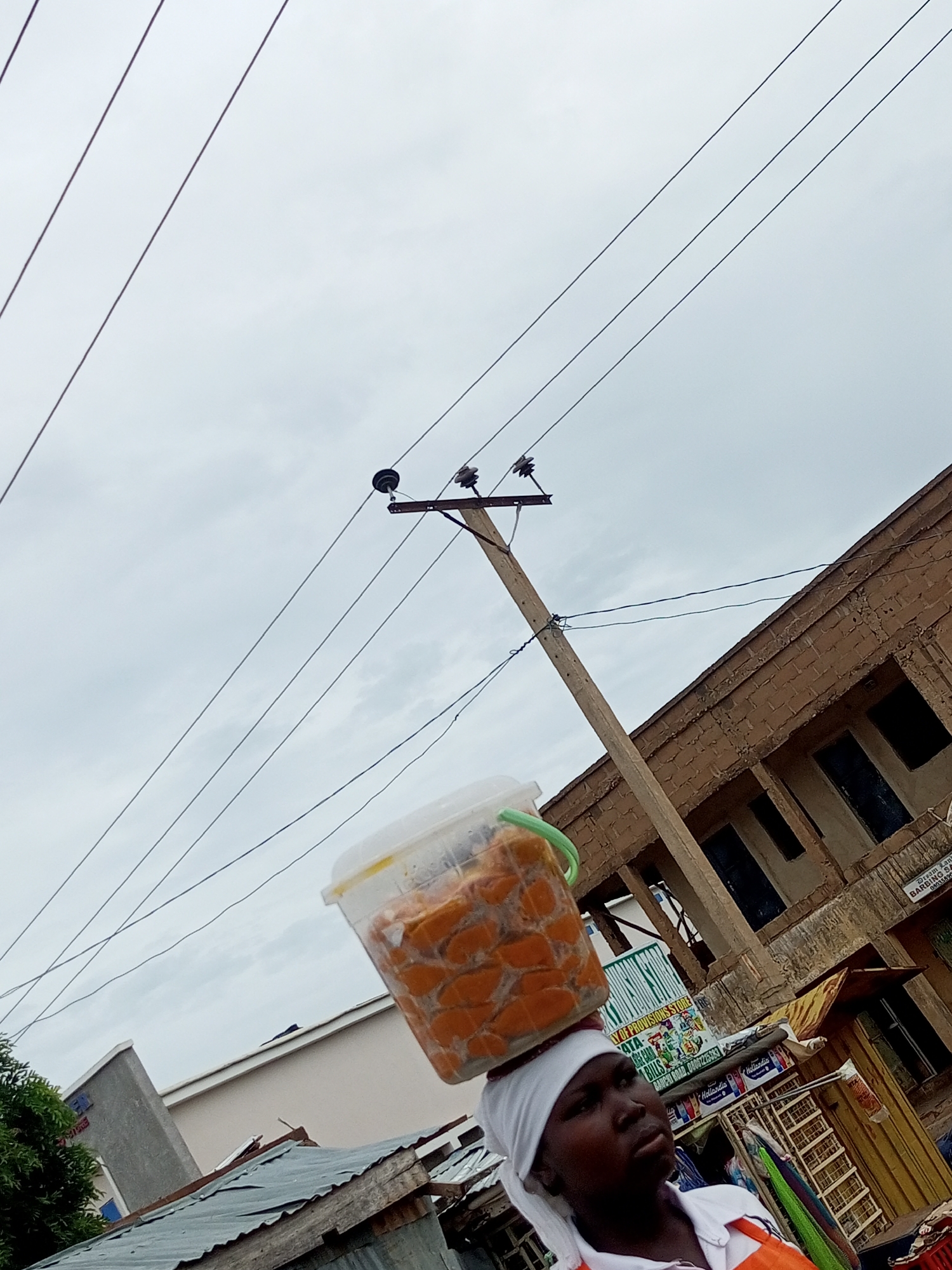 A woman balances a clear plastic tub filled with orange-colored food items on her head. She wears a white head covering and an orange and white shirt. In the background, a building with exposed brickwork is visible, along with a utility pole supporting multiple electrical wires. The scene is set outdoors, under an overcast sky, likely within a bustling urban or semi-urban environment in Tempure, Nigeria, given the context of street vendors and local architecture. Several stalls or shops are present, with some signage suggesting a marketplace. The overall impression is of a typical day in a Nigerian town, with individuals engaged in commerce and daily activities.