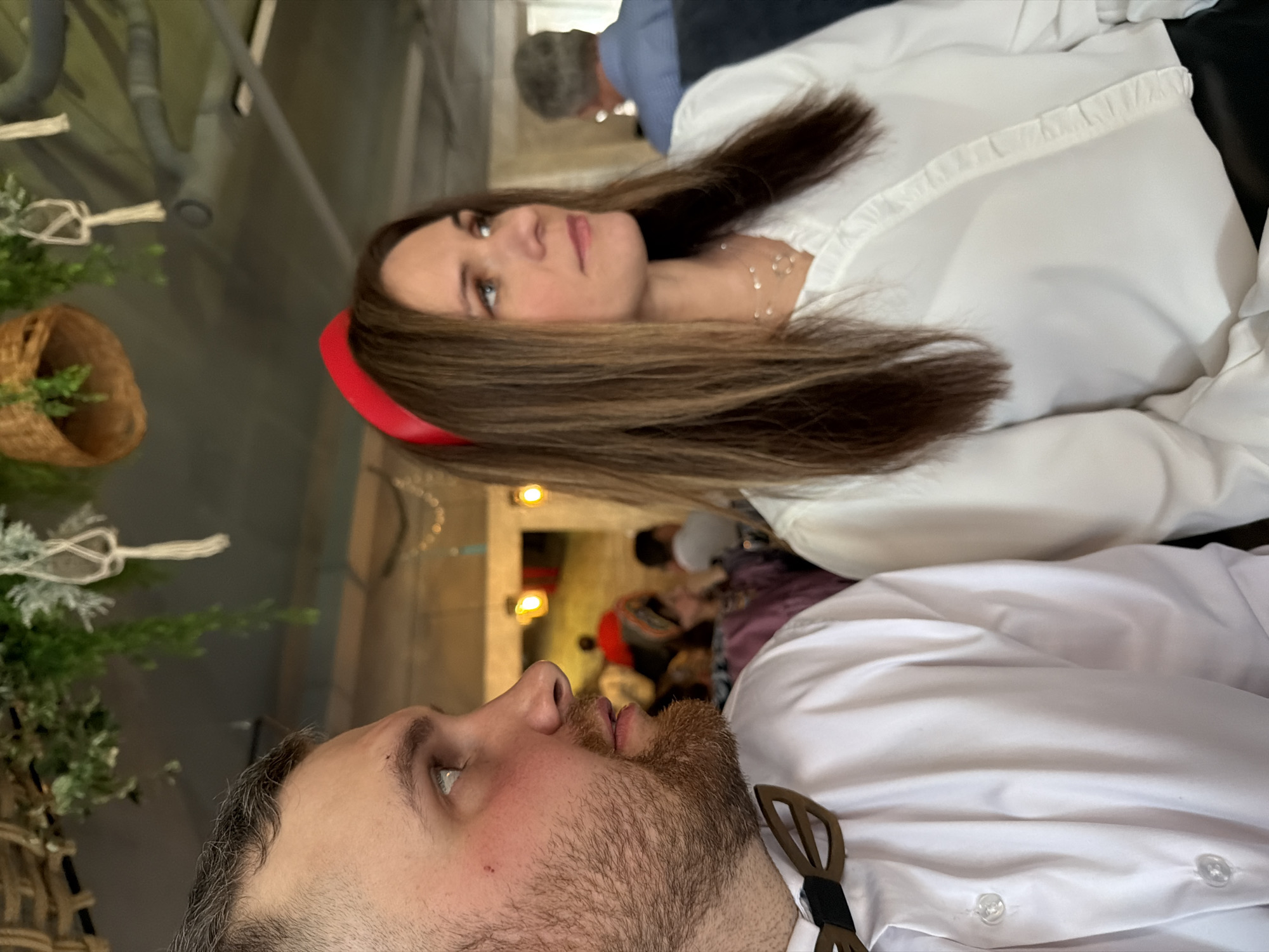 A man and a woman are in a close embrace. The man has a beard and a white collared shirt with a wooden bowtie. The woman has brown hair, a red headband, and a white collared shirt. The background shows foliage and blurred figures, suggesting a social gathering. The location is Chaikovsky, Russia.