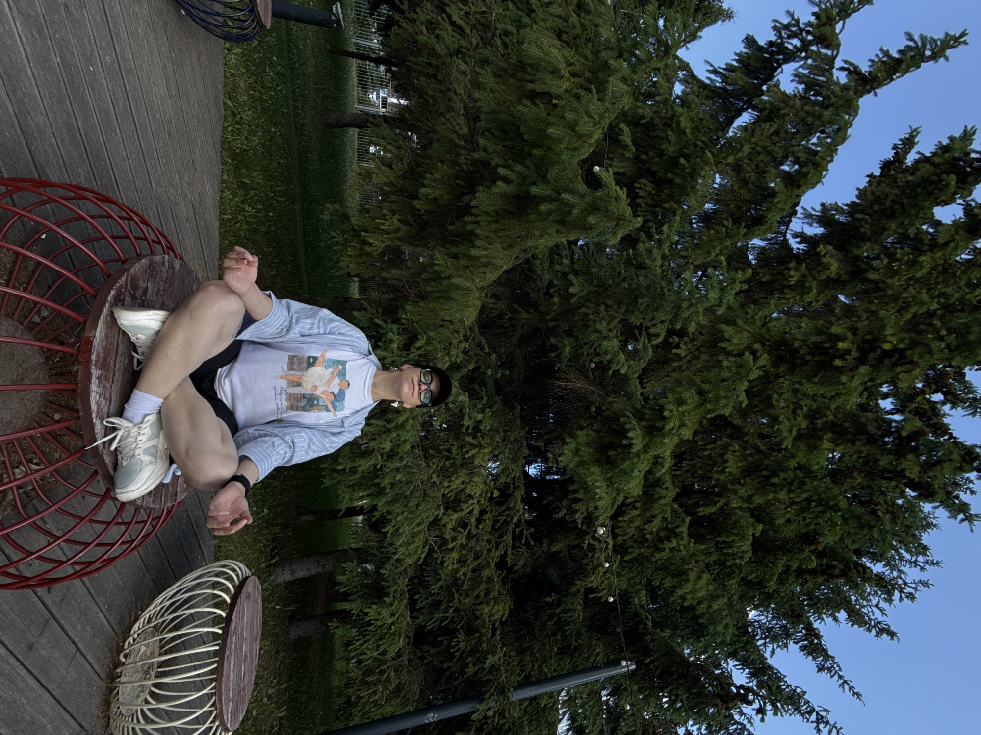 A person sits cross-legged on a red, wire-frame chair with a wooden top, located on a wooden deck. The person wears a light blue, button-down shirt over a white t-shirt with a design, black shorts, white socks, and light-colored sneakers. Glasses are perched on the person's face. A similar white wire-frame chair with a wooden top is visible near the red chair. Behind the person and the chairs is a large, green coniferous tree. The sky is visible in the upper right of the image and appears to be dusk. The location is Kazan, Russia.