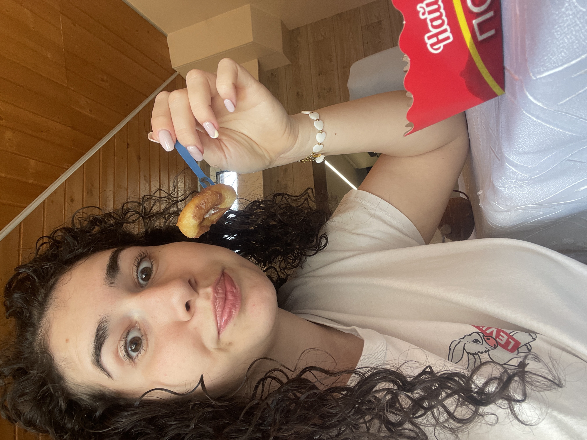 A young woman with curly brown hair is holding a donut with a blue plastic fork. She is wearing a white t-shirt and a white bracelet with gold accents and heart-shaped decorations. To her right is a red and yellow donut box. The background includes wooden paneling on the wall. The image does not provide enough information to identify the exact location in Sofia, Bulgaria.
