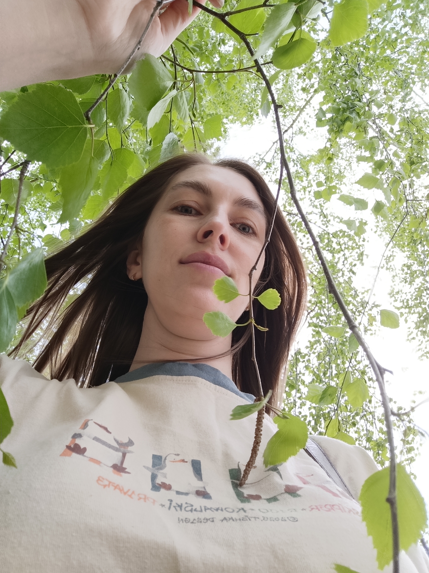 A low-angle shot depicts a woman with long brown hair standing under a tree with green leaves. The woman is centered in the frame, looking down at the camera. She is wearing a cream-colored t-shirt with a blue collar and a design featuring stylized images and text.
Above the woman, the leaves of the tree create a canopy, filtering the light. Branches are visible both in the foreground and background, some with small sprigs of leaves.
Given the prompt's request to determine the location as Chaikovsky, Russia, and the presence of birch-like trees, the environment is likely a park or green area in this city.