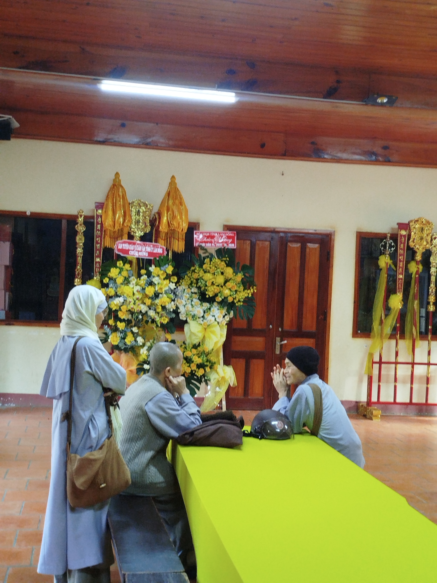 Three individuals are present in an indoor space that appears to be a temple or a community hall. The person on the left is wearing a white hijab and a light blue robe with a brown shoulder bag. A person sits at a table with their head in their hand and is wearing a grey vest and a blue shirt. The person on the right is wearing a black beanie and is leaning on the table with their hands in front of their face. A brown bag and a black helmet are on the table. The table is covered with bright green fabric. Behind the individuals is an arrangement of yellow flowers, as well as banners. Windows and a dark wooden door are also visible. The ceiling is made of dark wooden planks. The OCR information indicates that the image was taken in Da Lat, Vietnam.